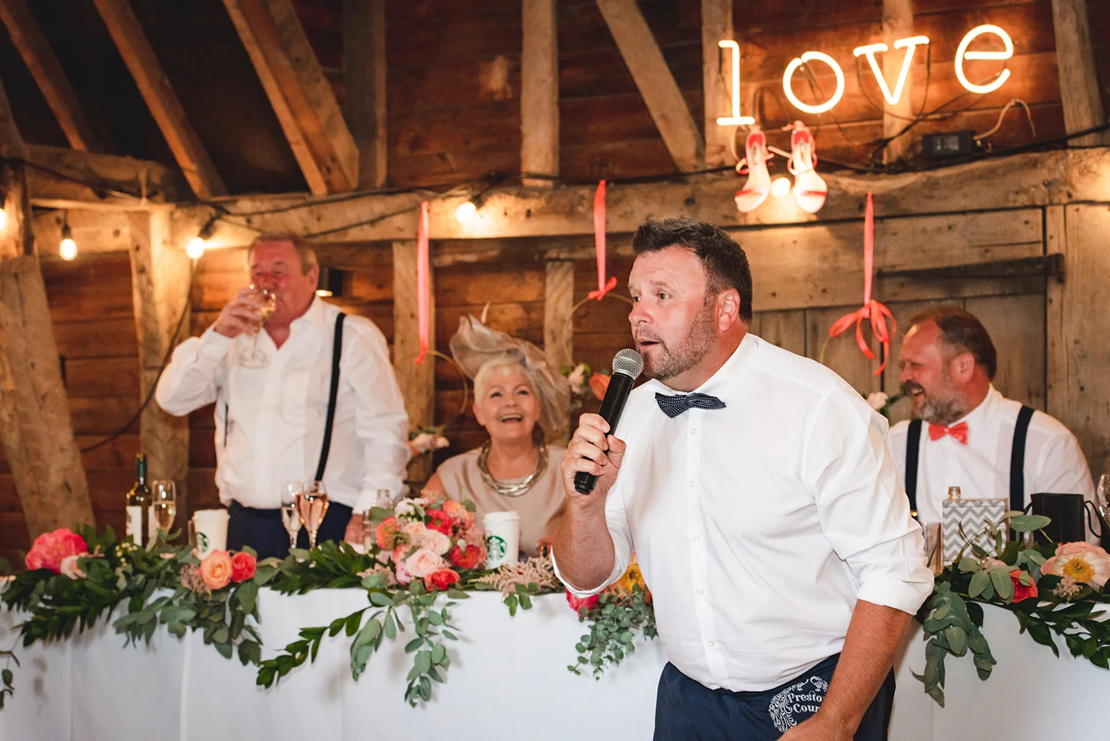 A guest delivers a speech with a microphone during a wedding reception inside a rustic oak-framed barn decorated with floral arrangements and a glowing neon sign.