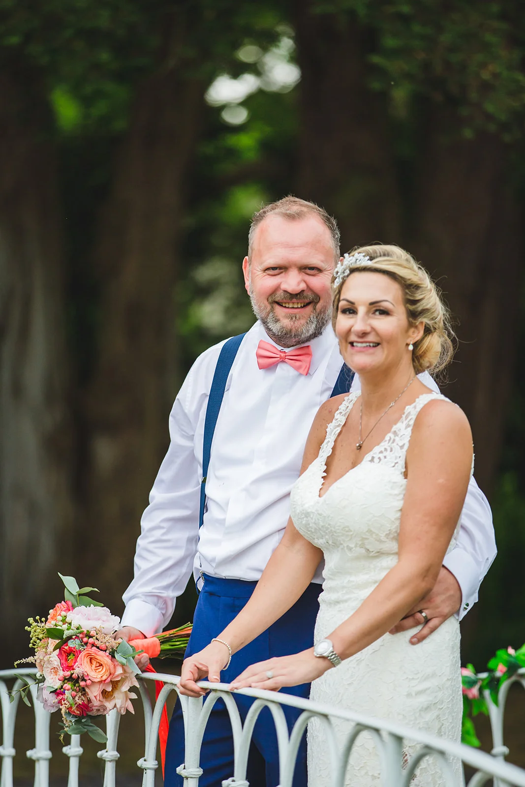 A smiling bride in a lace wedding dress and a groom in blue trousers and a pink bow tie stand together outdoors, holding a colourful bouquet.