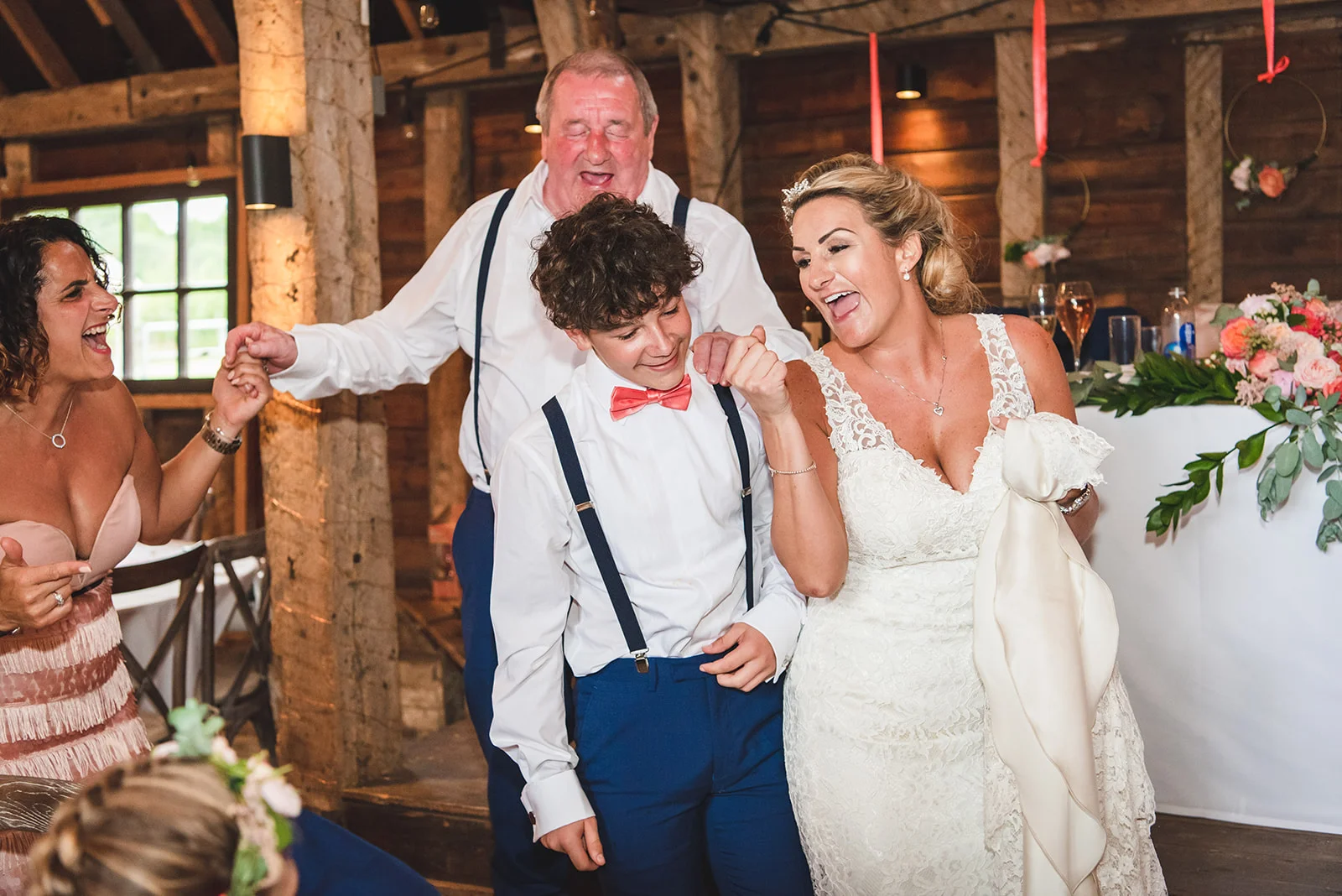 A laughing bride in a lace wedding dress celebrates joyfully inside a rustic timber barn alongside guests and a young boy in braces.