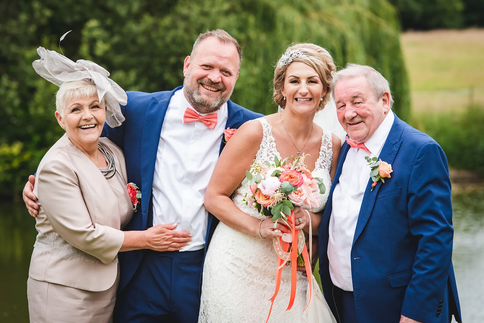 A smiling bride and groom pose happily outdoors alongside the mother and father of the bride, all dressed in formal wedding attire.