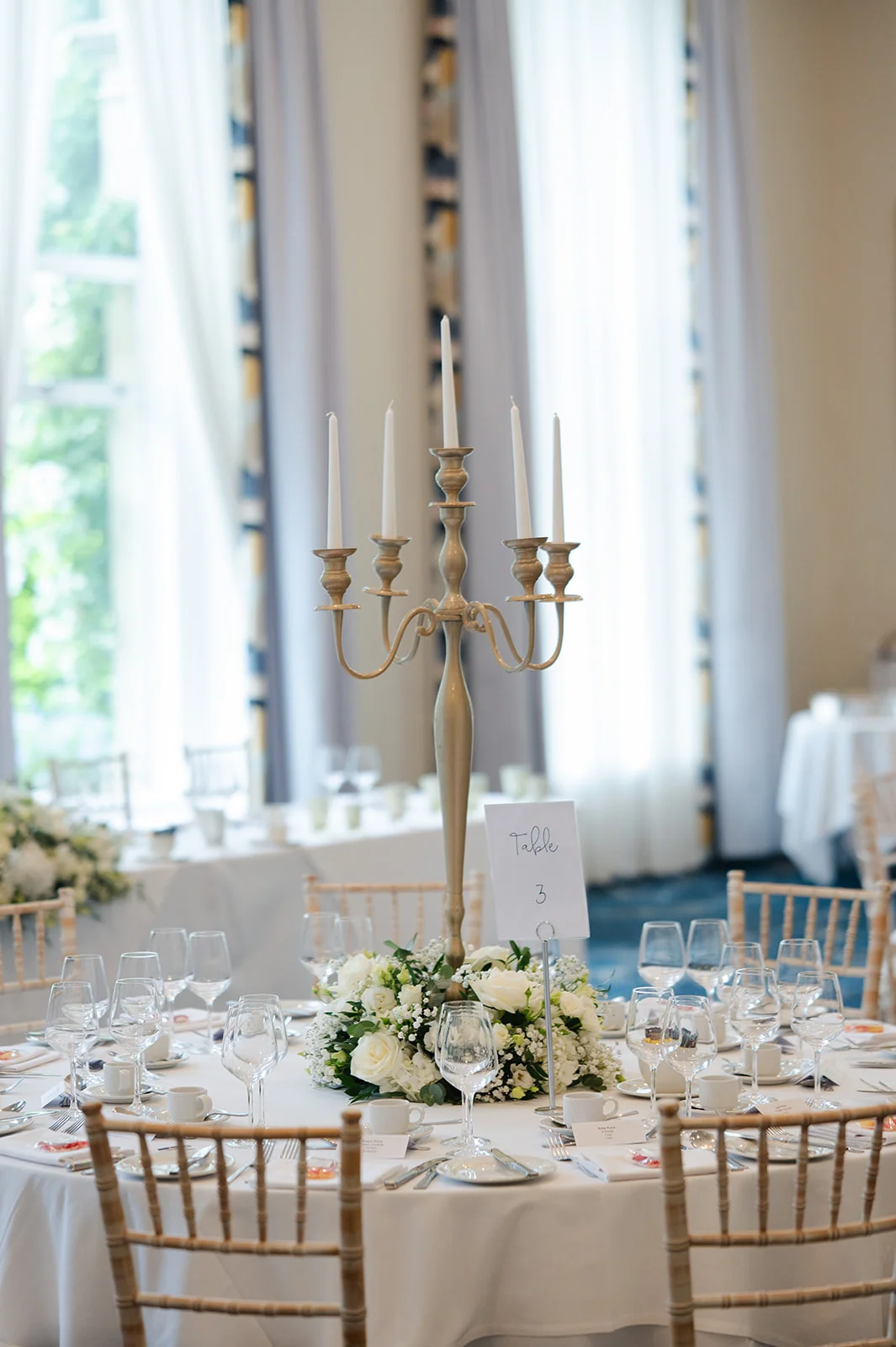 An elegant wedding tablescape featuring a tall silver candelabra centrepiece surrounded by white roses and lush greenery on a round table.