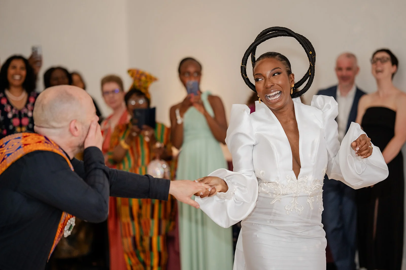 A joyful black bride wearing a structured white gown and a striking halo headpiece laughs ecpressionately while holding hands and dancing with her groom in front of a cheering crowd of wedding guests.
