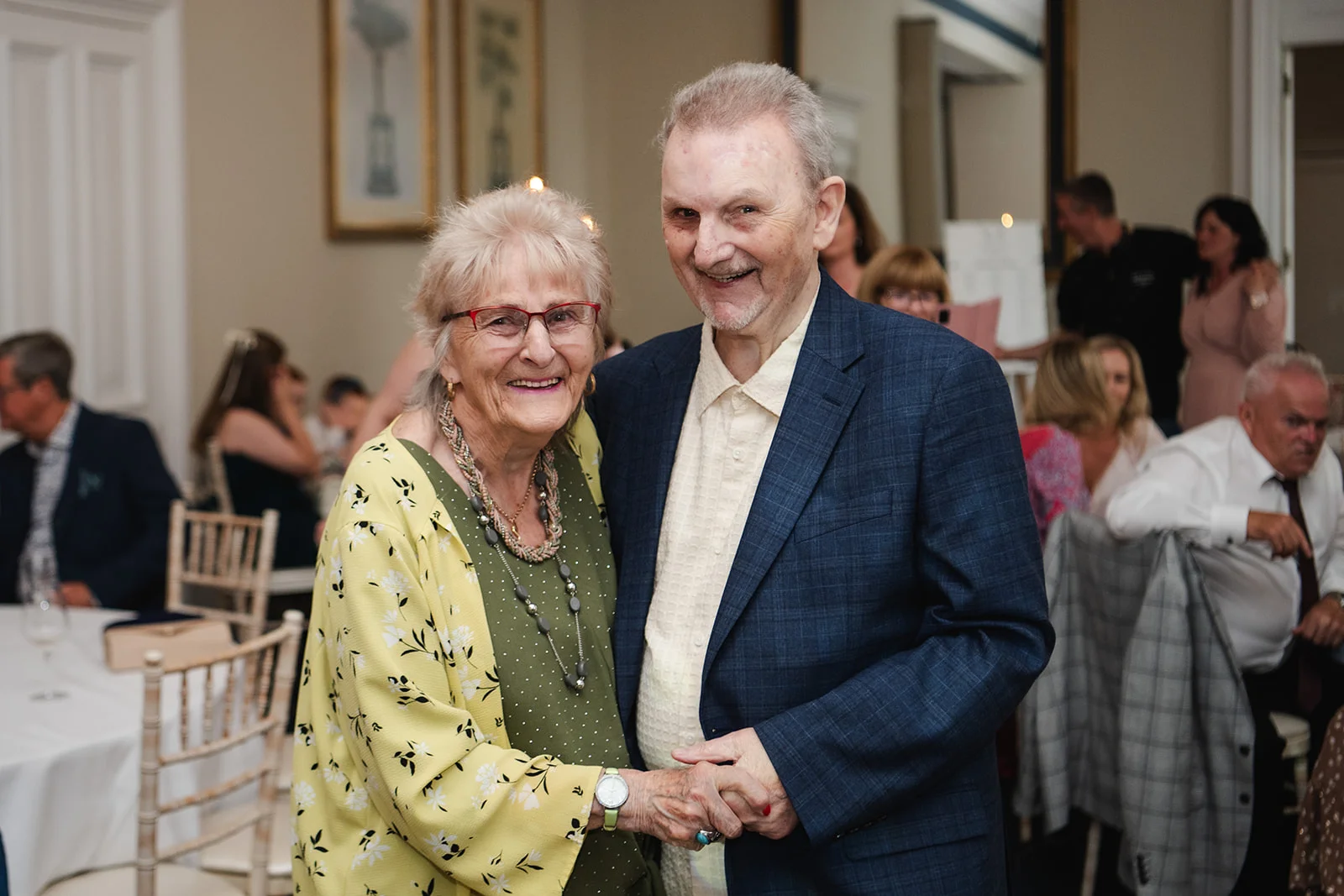 An elderly couple smiles warmly while holding hands during a joyful wedding reception indoors.