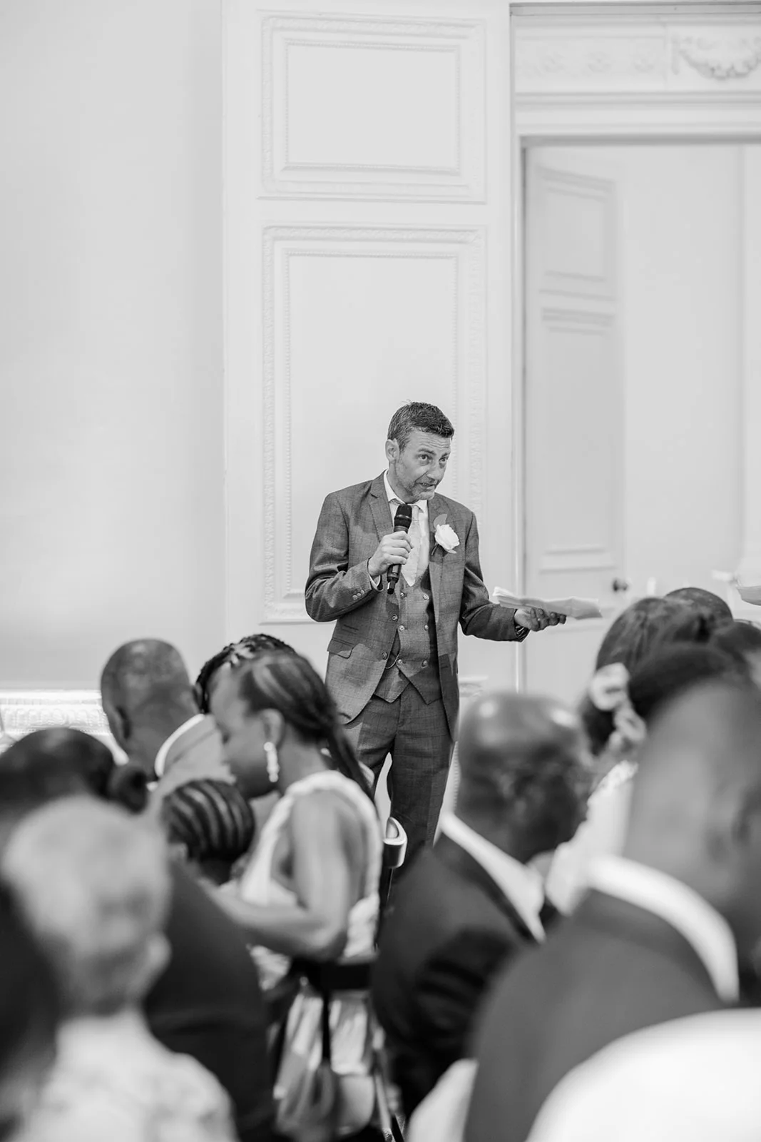 A groom in a three piece grey suit delivers a speech into a microphone, holding notes while standing before seated wedding guests in a bright, panelled room.