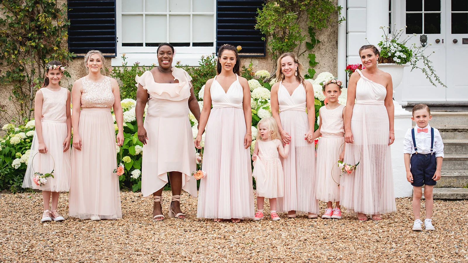 A diverse bridal party of bridesmaids, flower girls, and a page boy in pink attire pose together outside a country house.