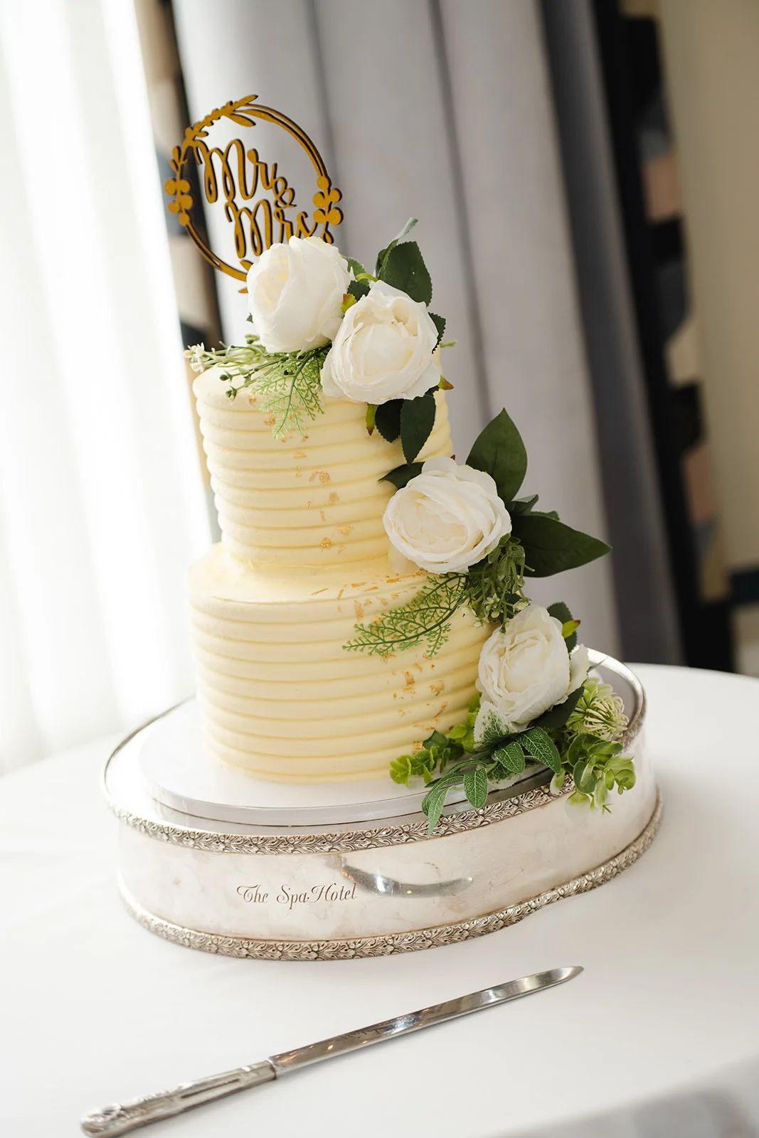 An elegant two-tier wedding cake with textured buttercream and gold leaf accents features a cascading floral arrangement of white roses and green foliage, topped with a custom gold 'Mr & Mrs' topper.