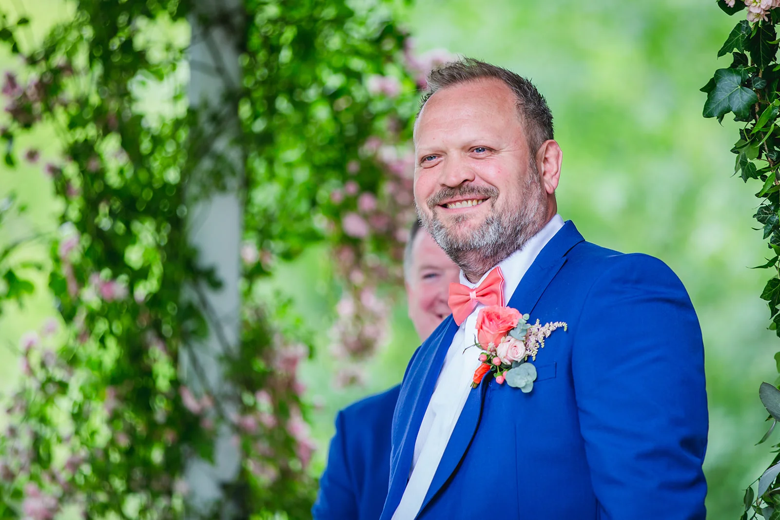 A smiling groom wearing a bright blue suit and a peach bow tie stands outdoors framed by lush green foliage and flowers.