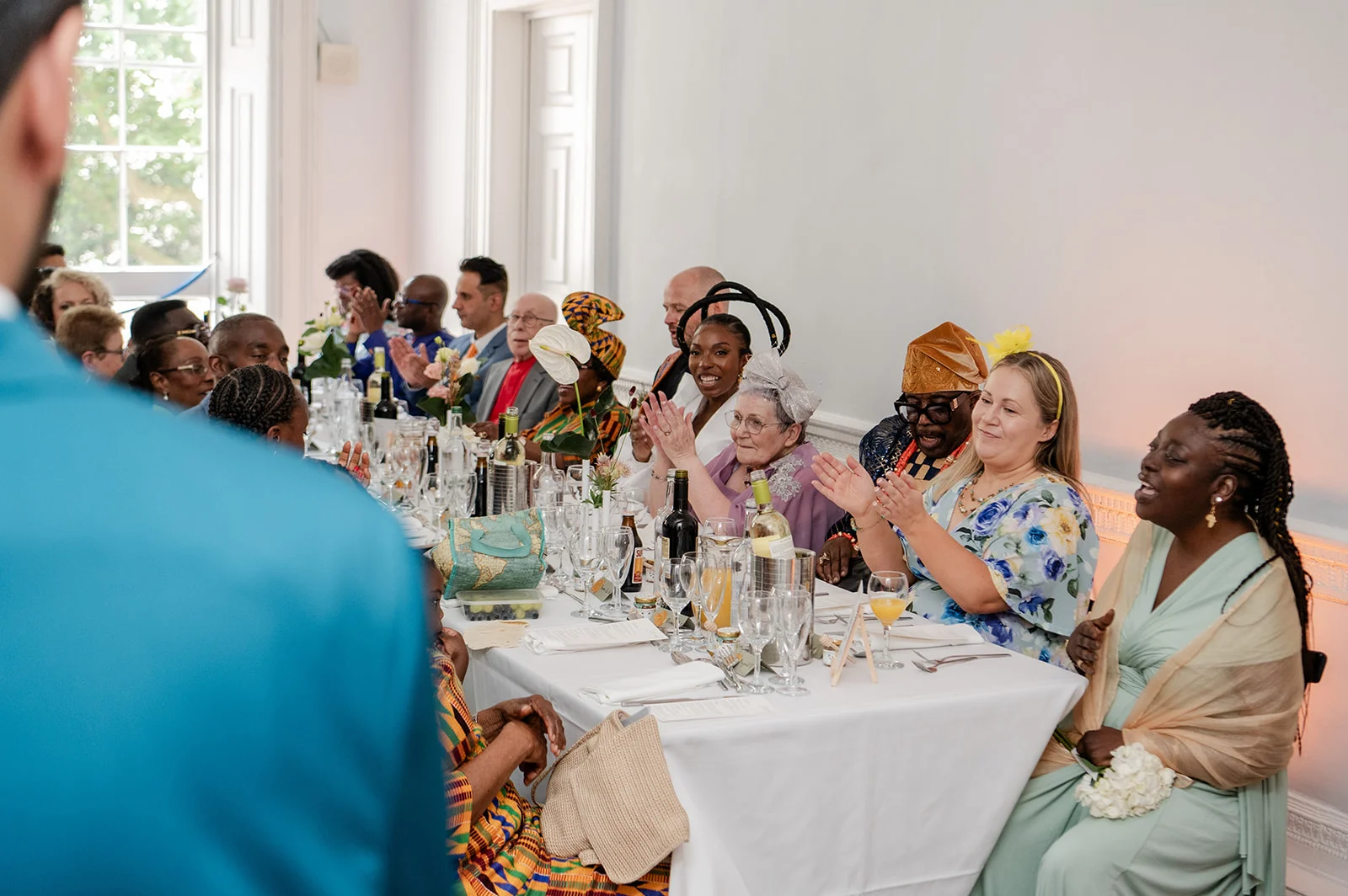 Wedding guests seated at a long banquet table laugh and clap during the reception speeches while a person in a bright blue jacket stands in the foreground.