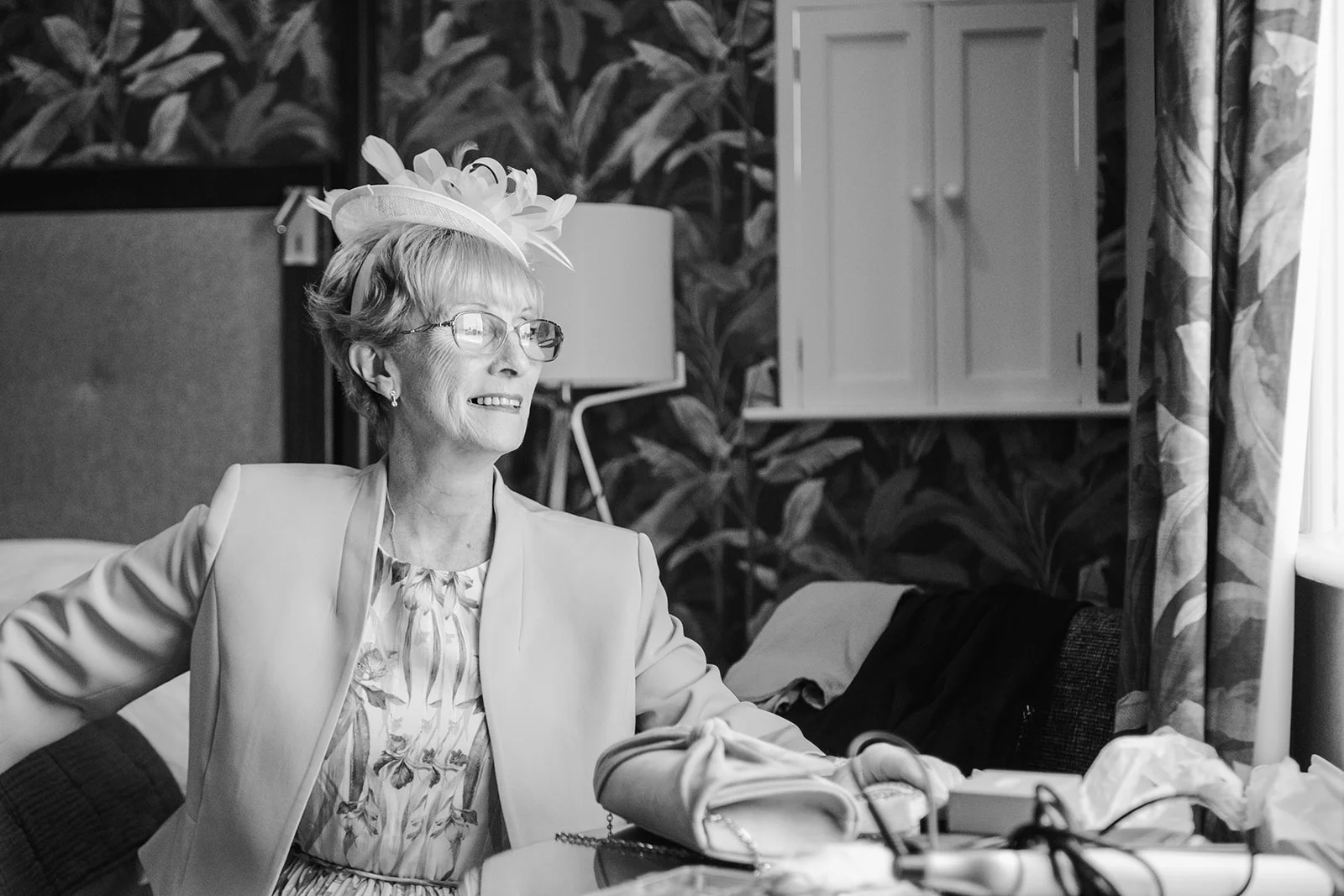 An elegant older guest wearing a patterned dress, structured jacket, and a chic fascinator smiles candidly in a warmly lit room.