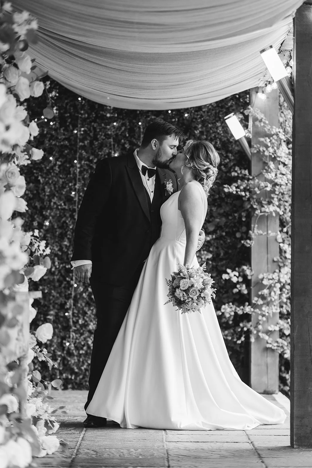 A newlywed couple share a tender kiss beneath a draped canopy and twinkling fairy lights while holding a floral bouquet.