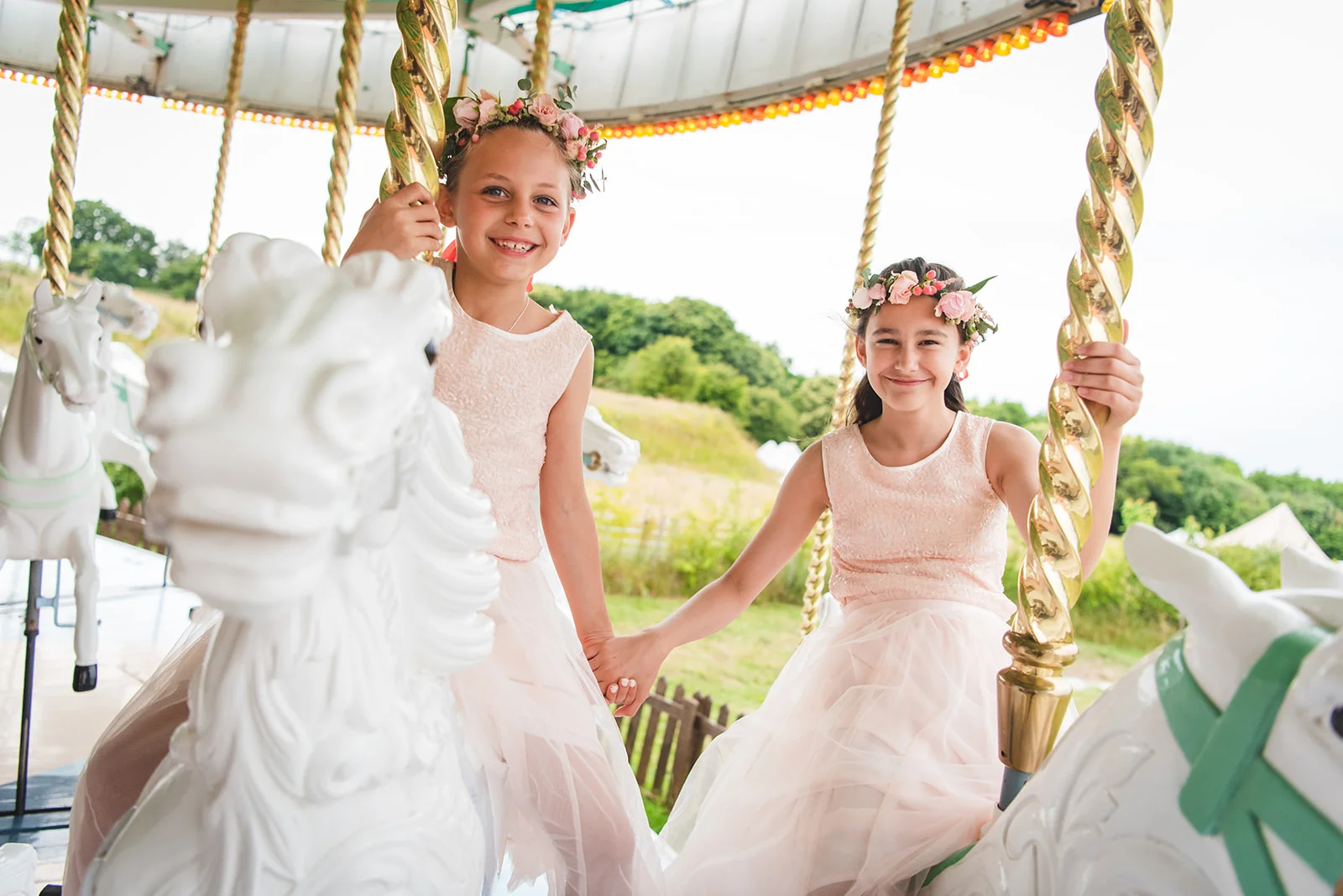 Two smiling junior bridesmaids wearing matching pink dresses and fresh flower crowns hold hands while riding a vintage carousel at a wedding reception.