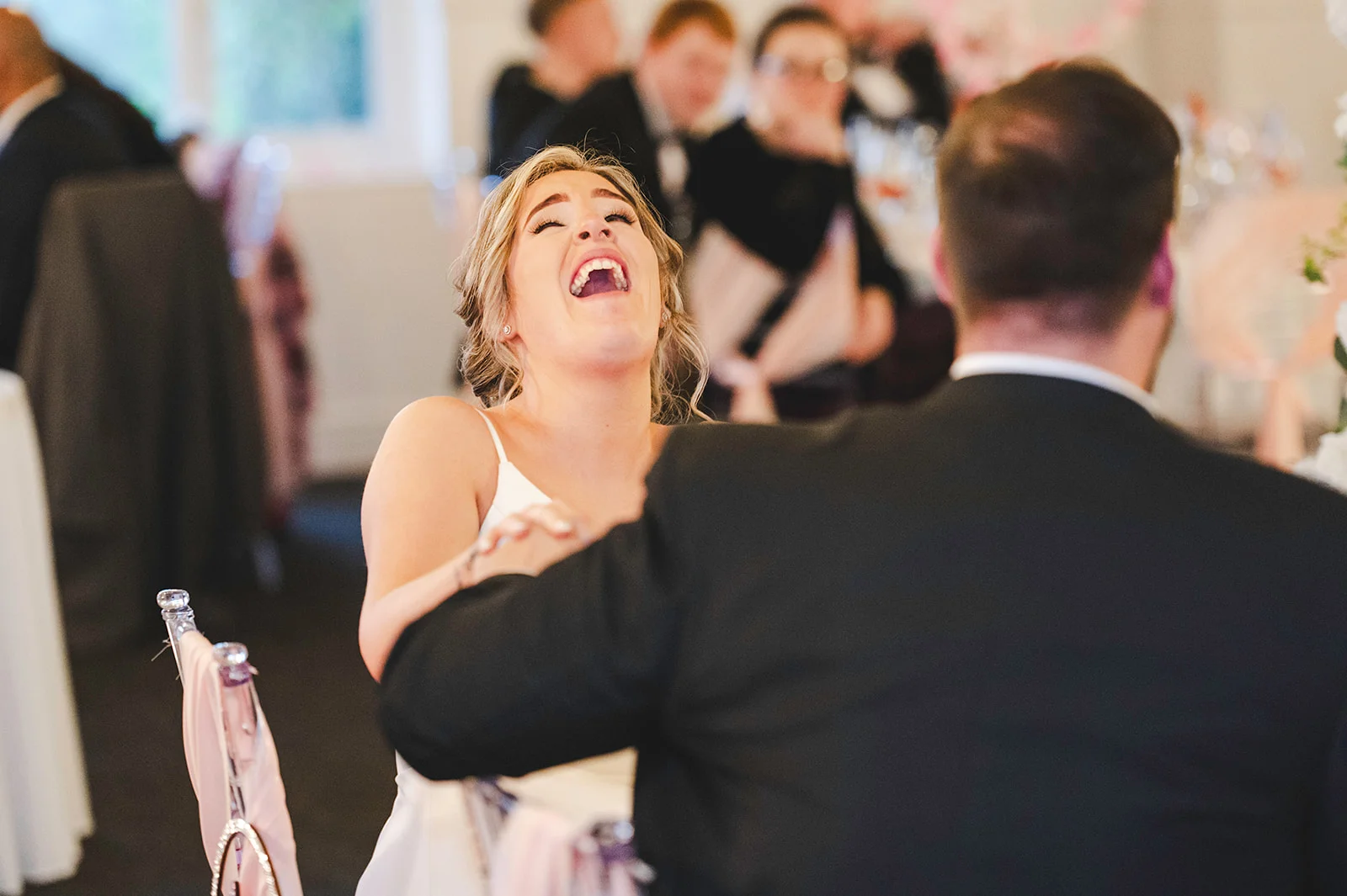 A newlywed bride throws her head back in roaring laughter during the wedding speeches, sitting beside her groom at the top table.