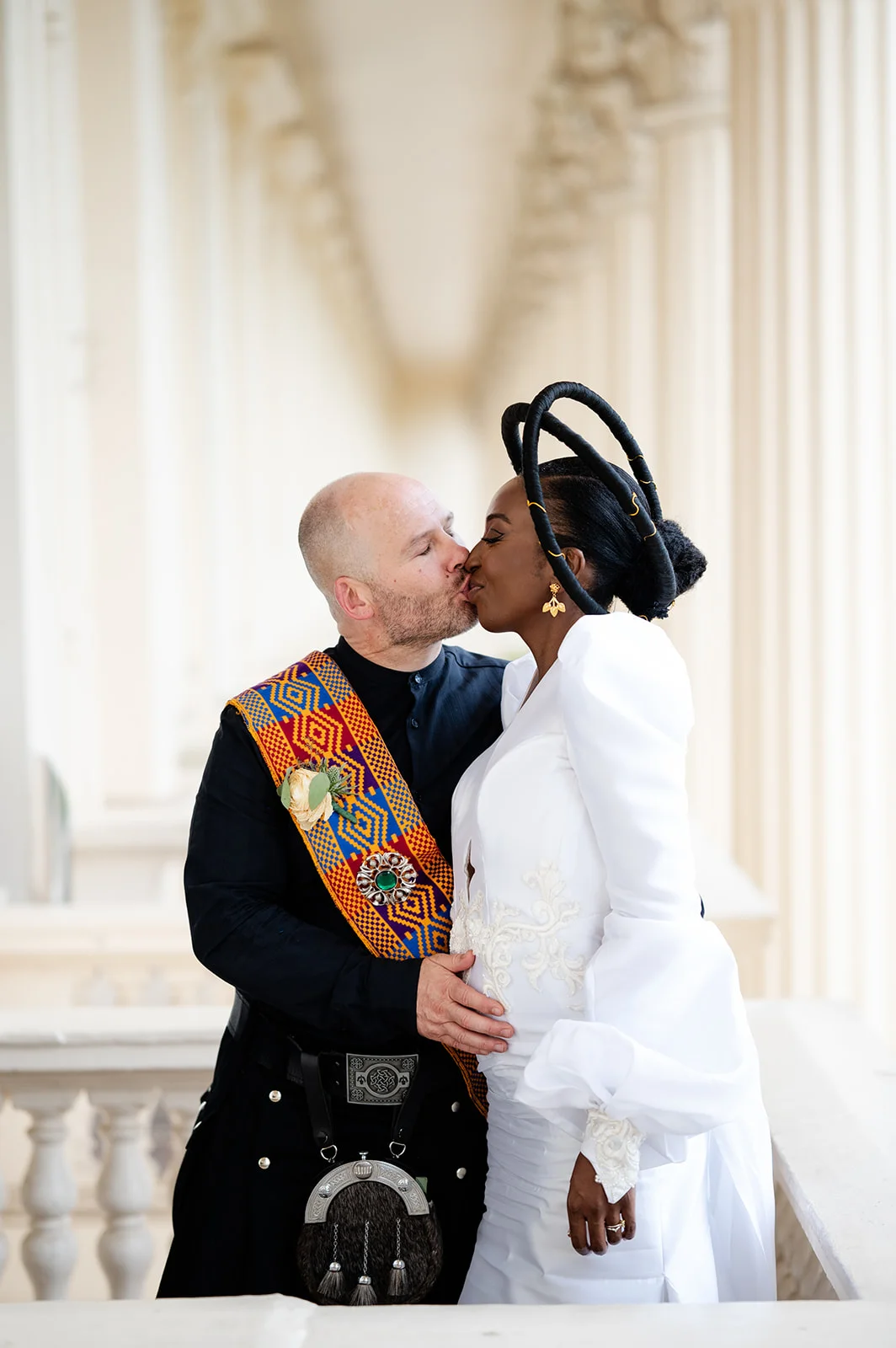 An interracial newlywed couple share a tender kiss, with the groom wearing a black kilt and colourful tartan sash, and the bride wearing a white gown with statement sleeves and an elaborate sculptural headpiece.