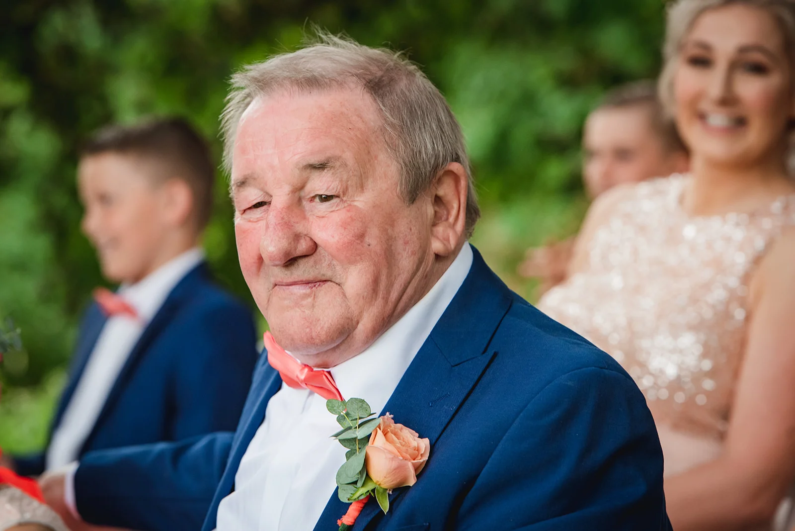 An elderly grandfather wearing a navy suit and coral bow tie smiles candidly during a wedding celebration.