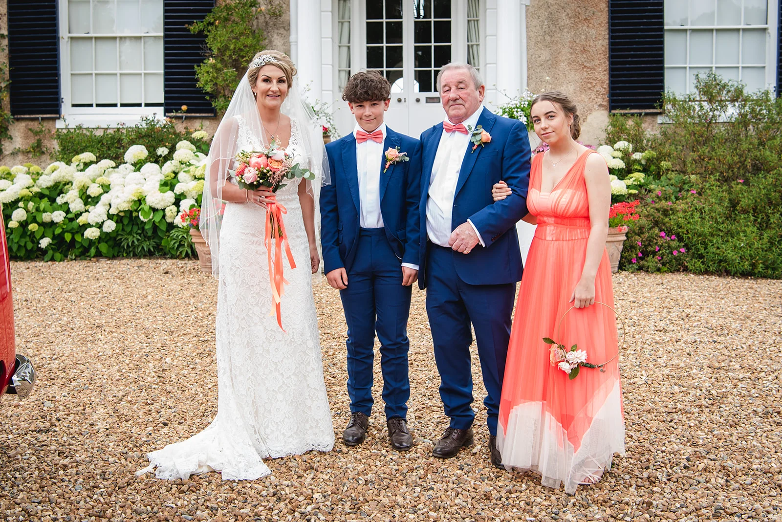 A formal family wedding portrait featuring a bride in a lace gown, a young boy and an older man in blue suits with pink bow ties, and a bridesmaid in a vibrant coral dress outside a country house.