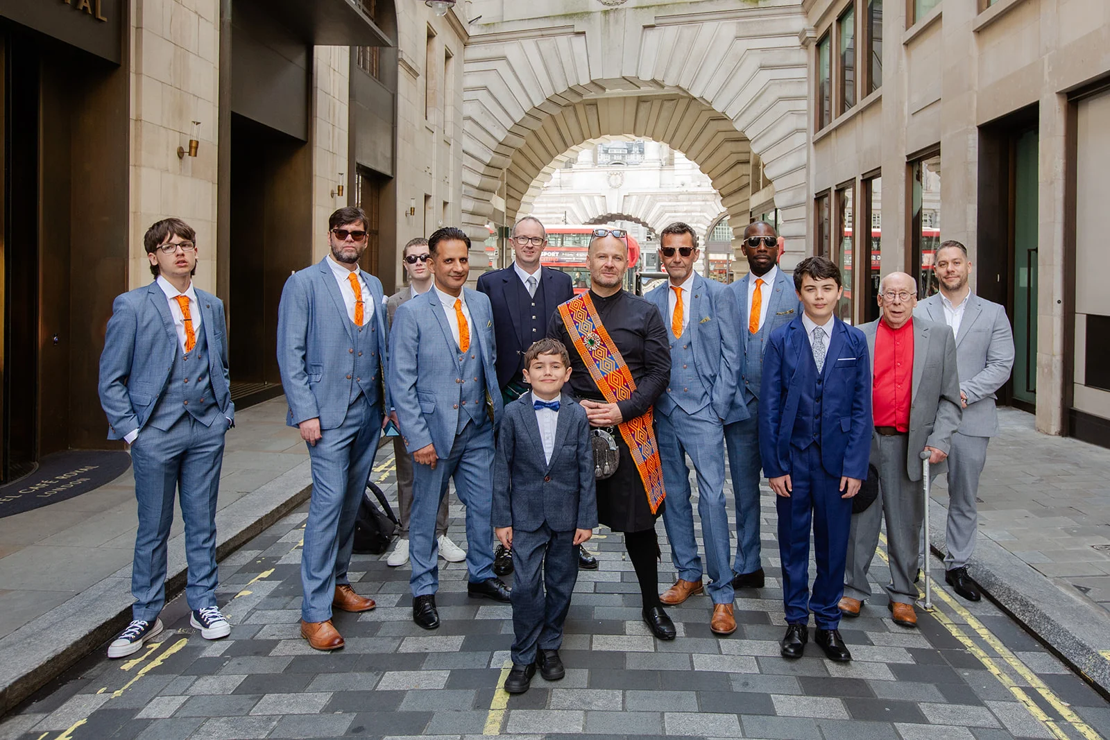 A stylish group of groomsmen, family members and a page boy pose together on a London street framed by stone architecture.