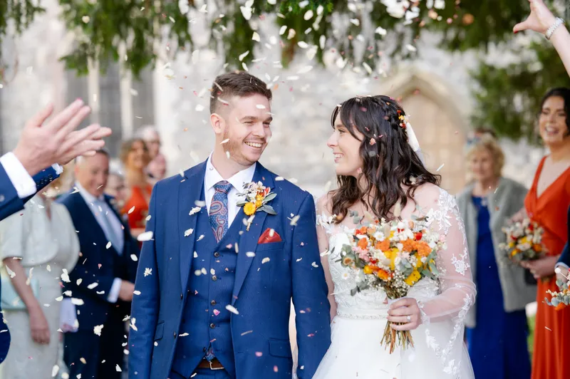 A smiling bride and groom walk through a shower of confetti outside a church, surrounded by cheering wedding guests.
