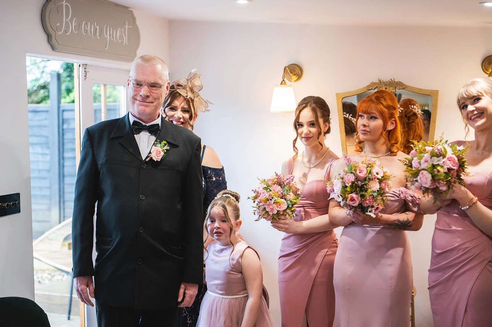 A proud father of the bride stands alongside bridesmaids in pink dresses holding floral bouquets and a tearful flower girl during wedding preparations.