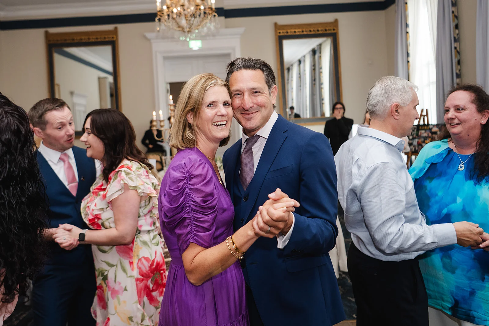 An older couple joyfully dances together during a wedding reception, surrounded by other guests in a brightly lit ballroom with ornate mirrors.