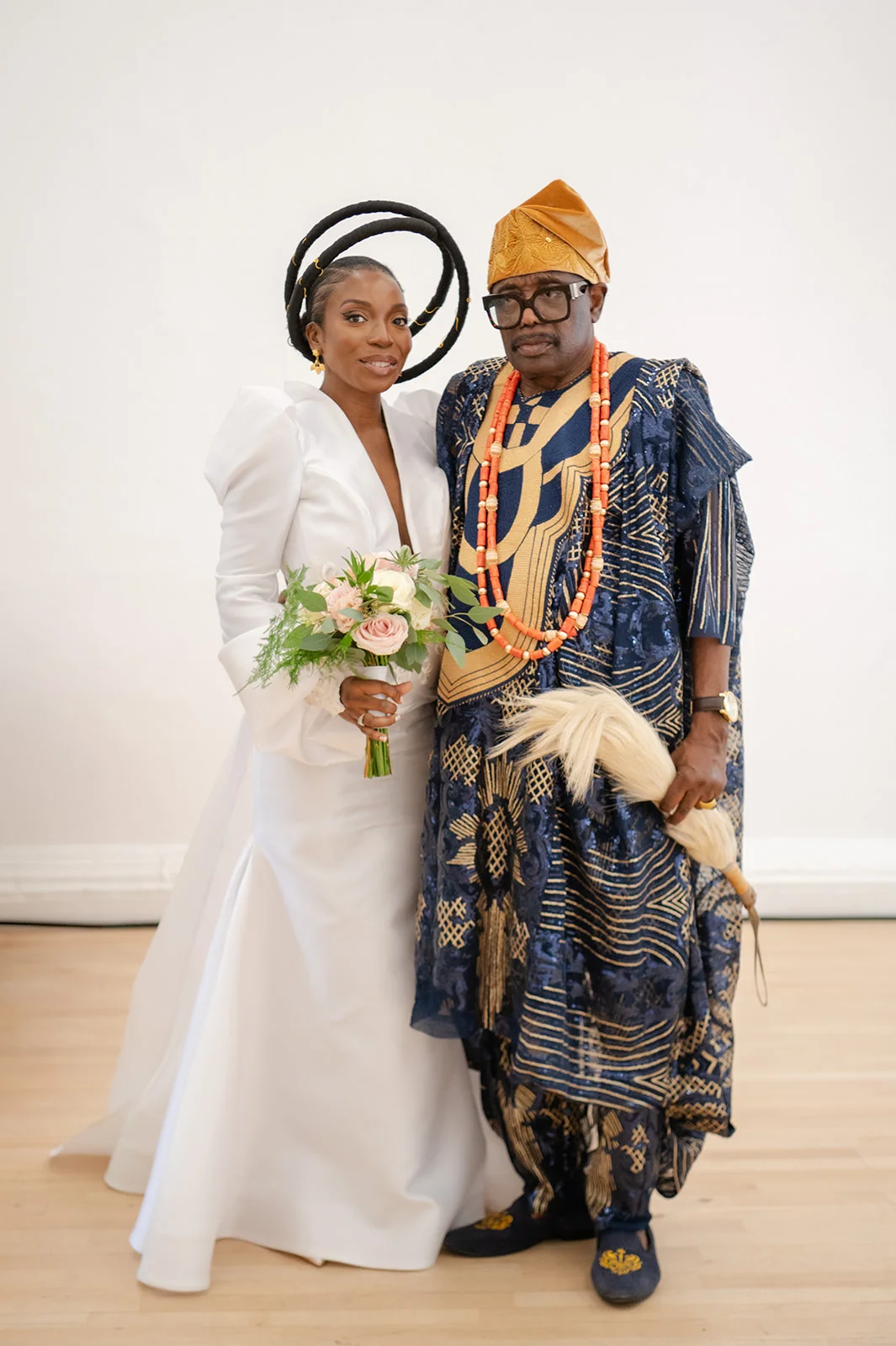 A newlywed couple poses together indoors, with the bride wearing a modern white gown and sculptural headpiece holding a bouquet, and the groom in a patterned navy agbada with coral beads.
