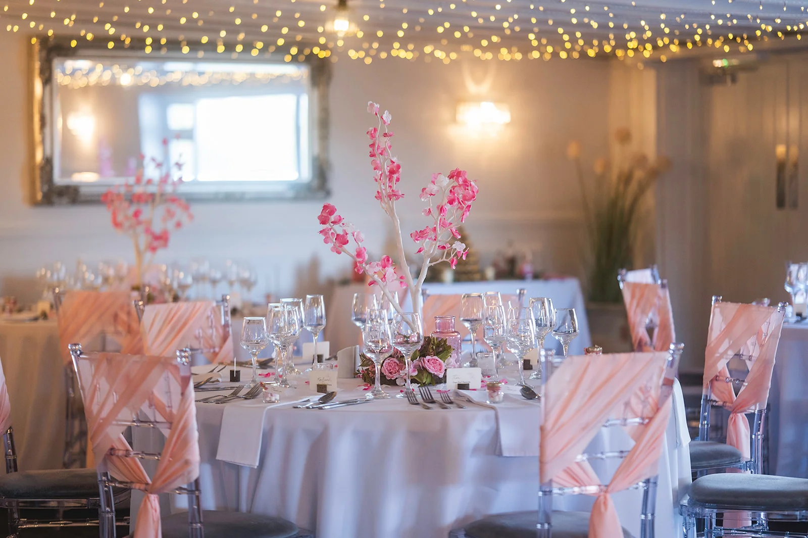 An elegantly decorated wedding reception room featuring round tables set with white linens, crystal glassware, and tall pink blossom branch centrepieces beneath a sparkling fairy light canopy.