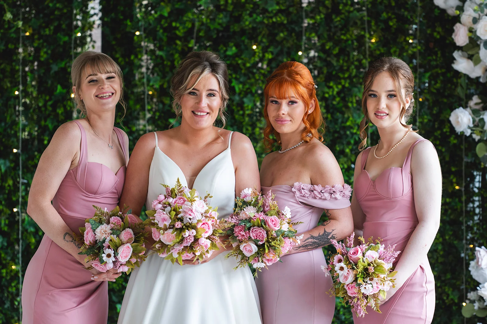A smiling bride and her bridesmaids in matching pink dresses pose together holding floral bouquets in front of a lush green wall.