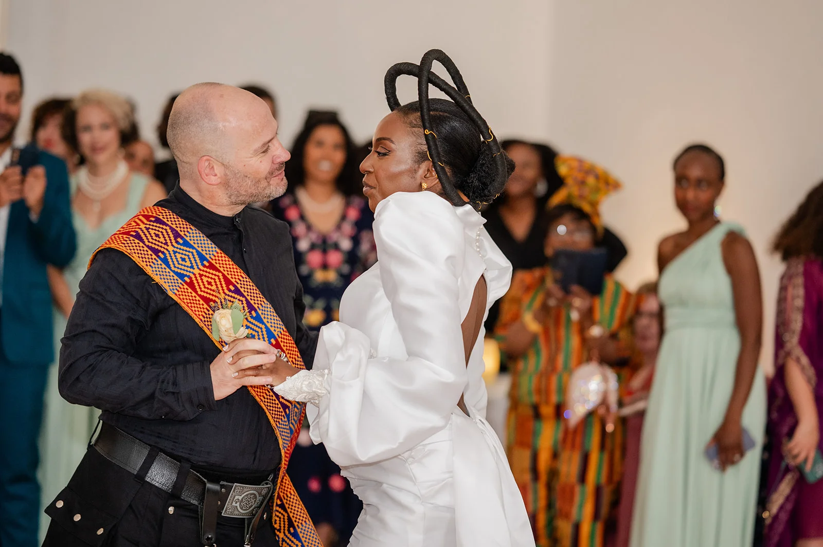 An interracial newlywed couple shares a tender first dance surrounded by family and guests, with the bride wearing a striking sculptural headpiece and white gown.