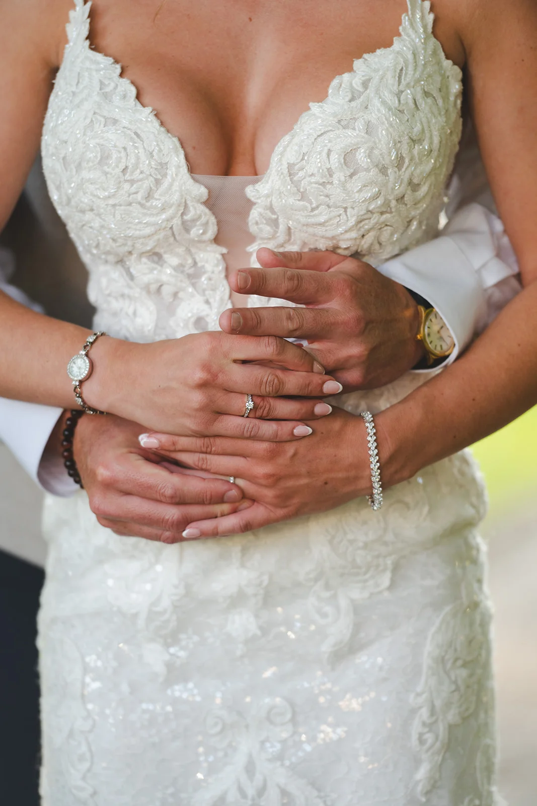 A close-up shot of a bride and groom embracing, showcasing their intertwined hands, wedding rings, and the intricate lace detail of the bridal gown.