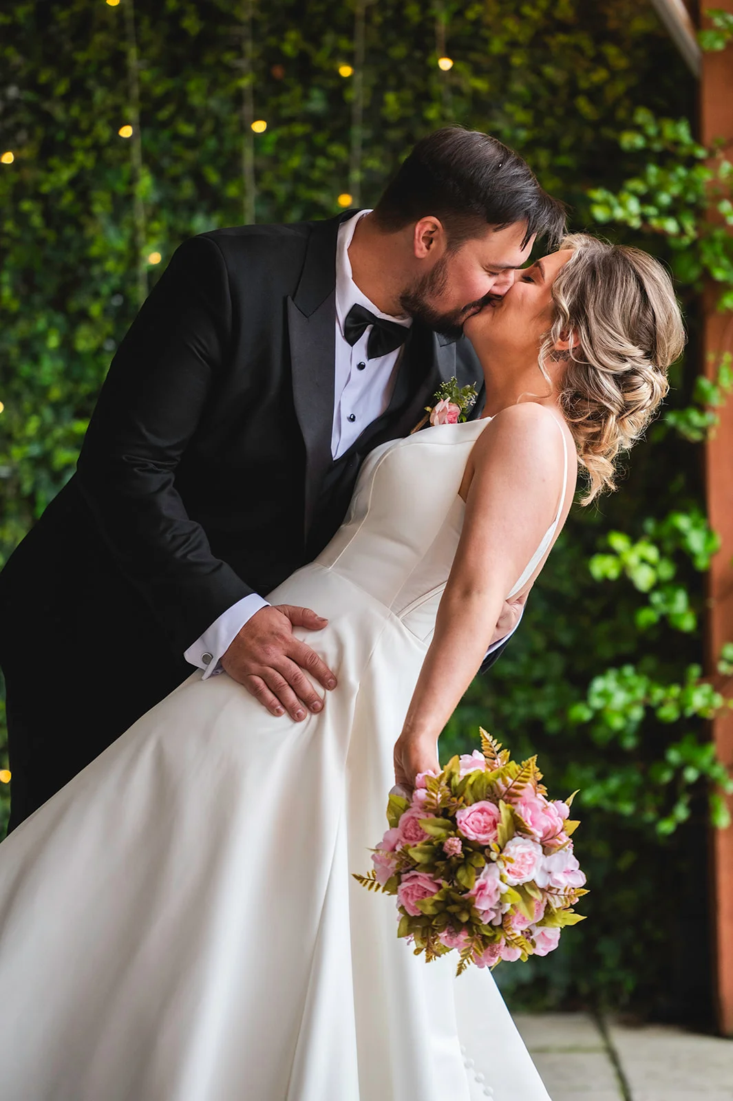 A groom in a classic black tuxedo dips and kisses his bride, who wears a structured white satin gown and holds a pink bouquet against a leafy green backdrop.
