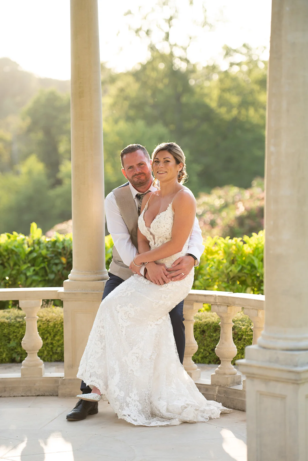 A newlywed couple embraces inside a stone gazebo during golden hour, with the bride seated gracefully in her lace a-line wedding dress.