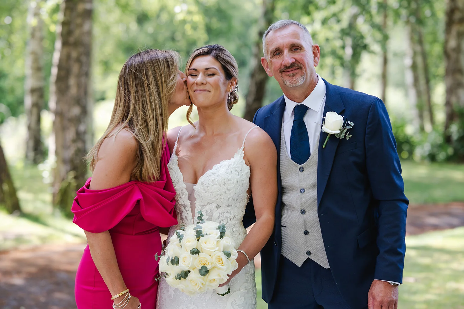 A mother kisses the smiling bride on the cheek outdoors in a woodland setting while the father of the bride stands proudly beside them.