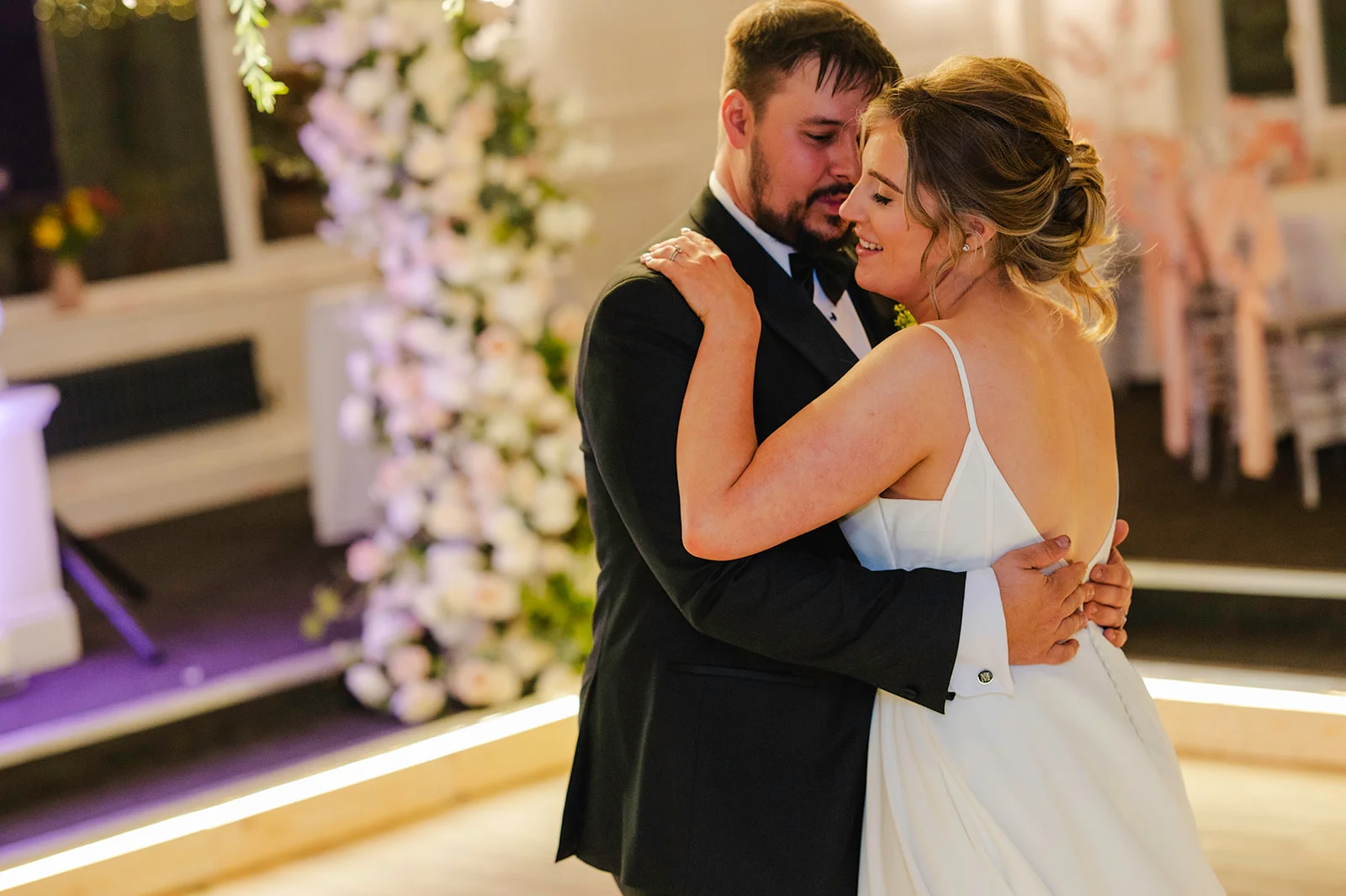 A newlywed couple share an intimate embrace during their first dance, with the bride wearing a sleek white gown and the groom in a classic black tuxedo.