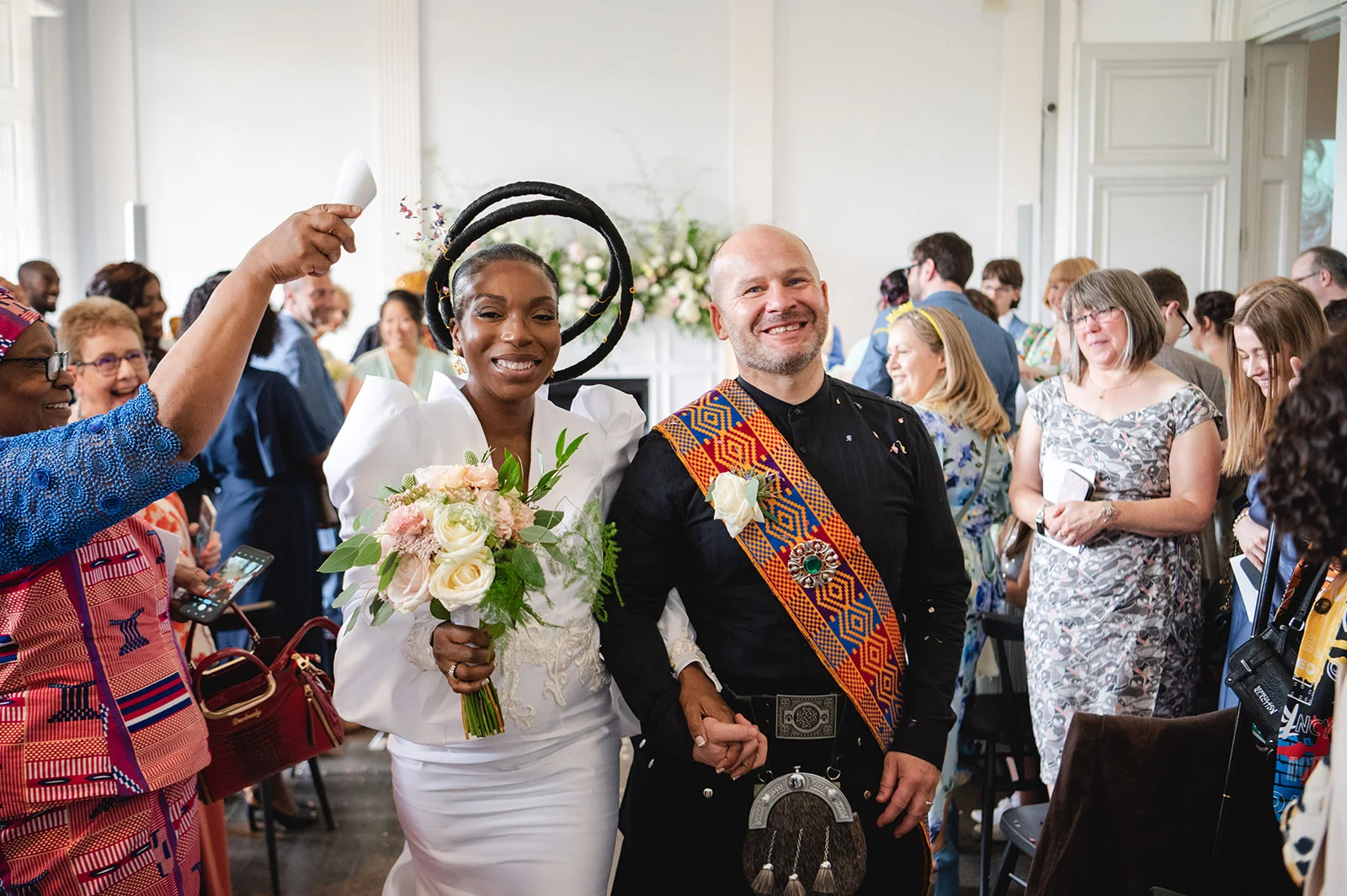 A joyful newlywed couple walks down the aisle hand in hand, with the bride wearing a white gown and dramatic sculptural headpiece while holding a bouquet, and the groom wearing a traditional kilt with a patterned sash.
