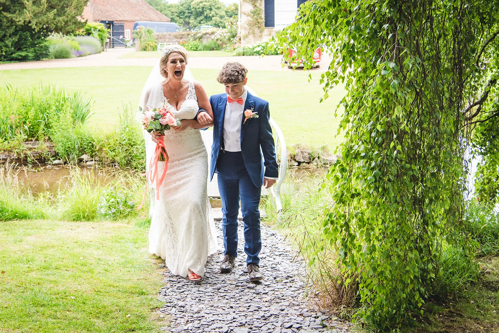 A joyful bride in a lace wedding dress holding a bouquet walks along a garden path next to a young boy in a blue suit.