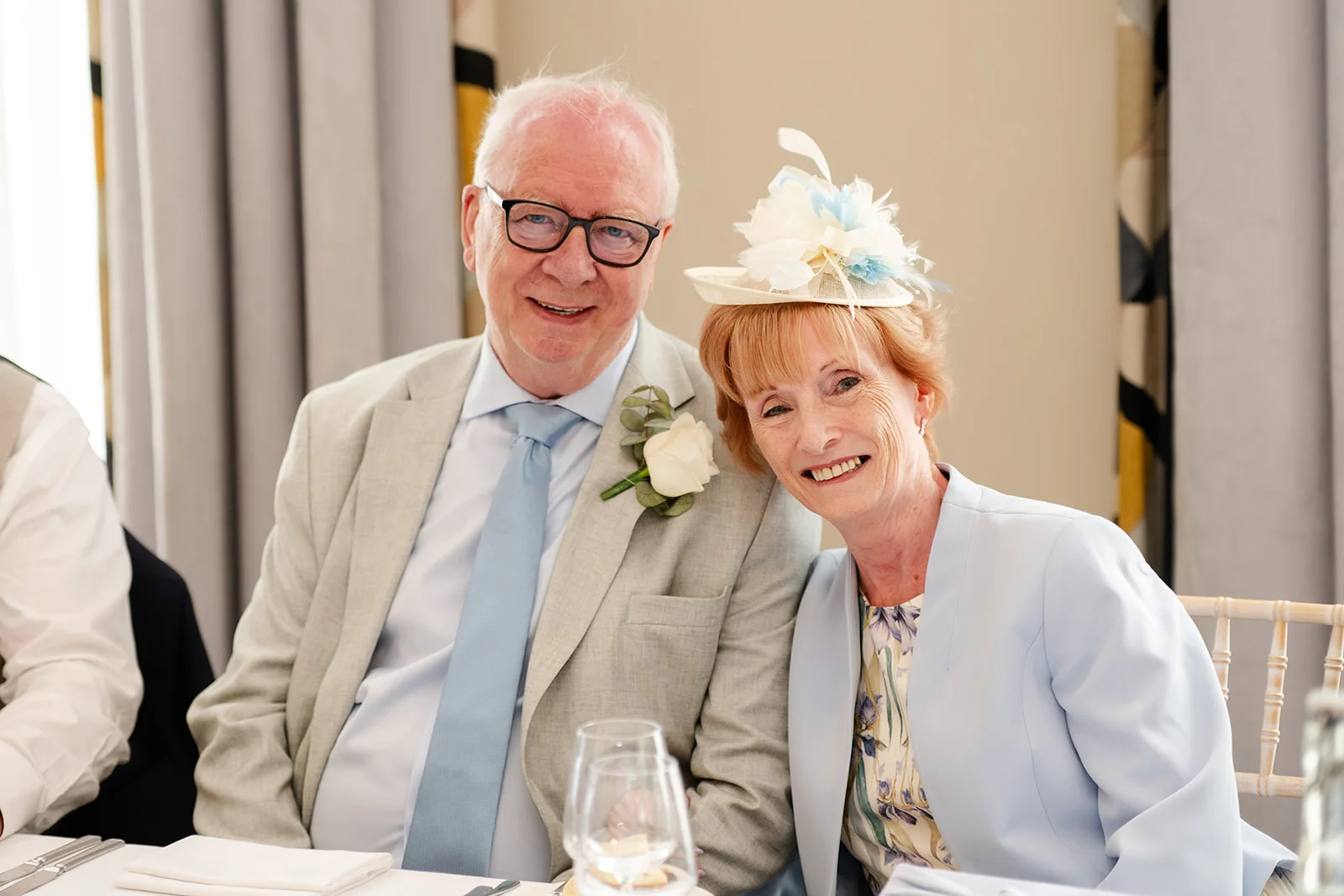 An older couple smiling happily together at a wedding reception, with the gentleman wearing a light grey suit and blue tie and the lady wearing a pale blue jacket and matching fascinator.