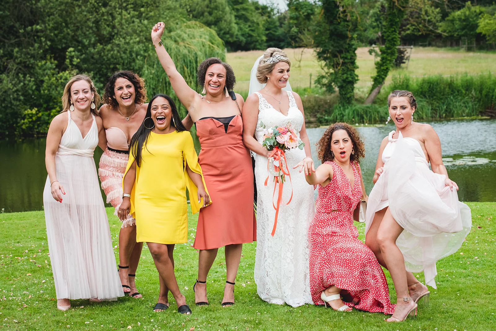 A joyful bride in a lace gown and tiara poses outdoors by a lake, surrounded by cheering and laughing bridesmaids wearing mismatched colourful dresses.