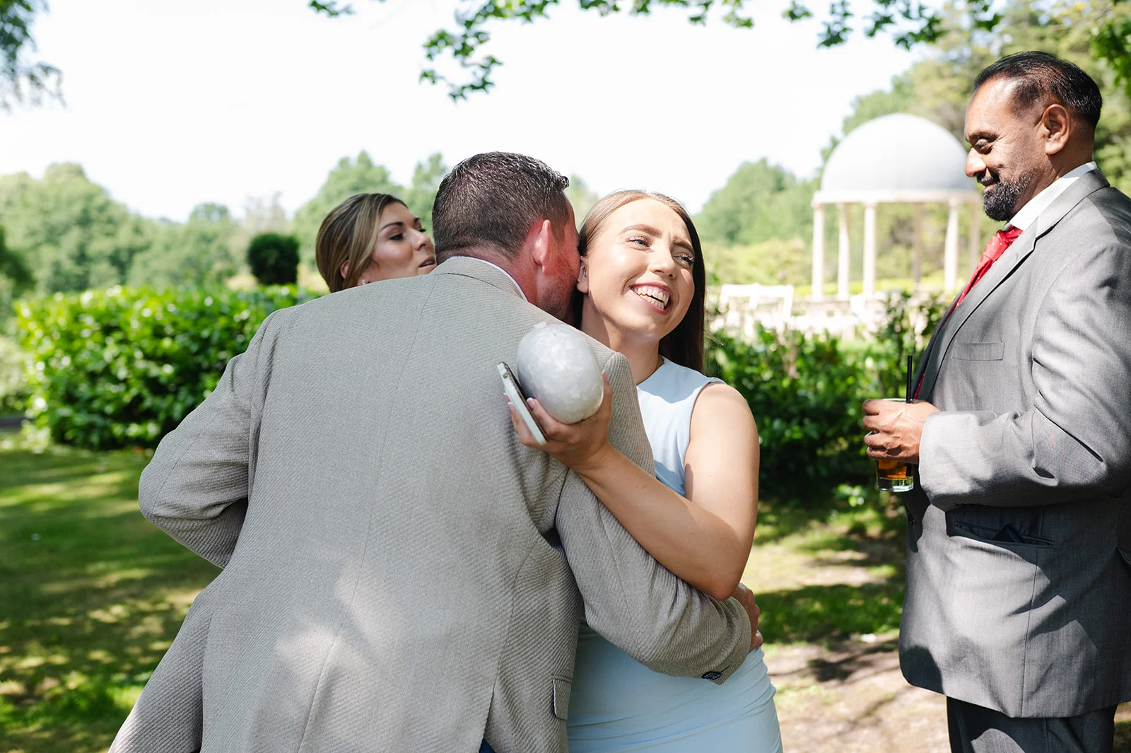 A guest in a light suit embraces a smiling woman in a pale blue dress outdoors at a summer wedding reception.
