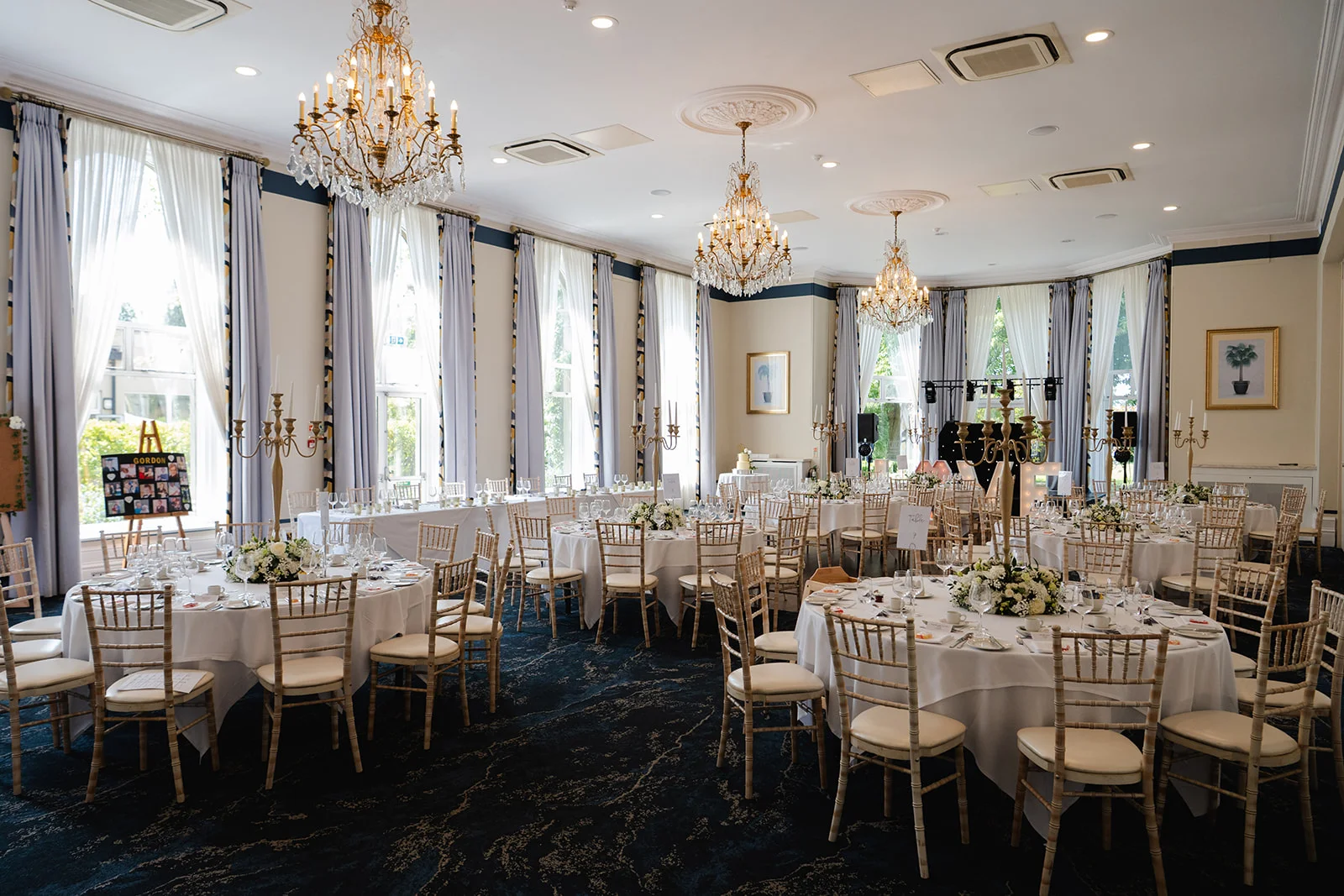 An elegant hotel ballroom set up for a wedding breakfast, featuring round tables with white linens, gold Chivarti chairs, tall candelabras, and crystal chandeliers.