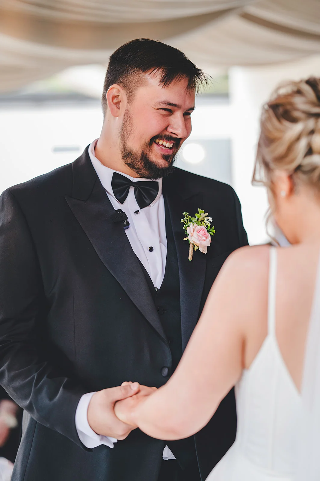 A smiling groom in a black tuxedo and bow tie holding hands with his bride during their wedding ceremony.