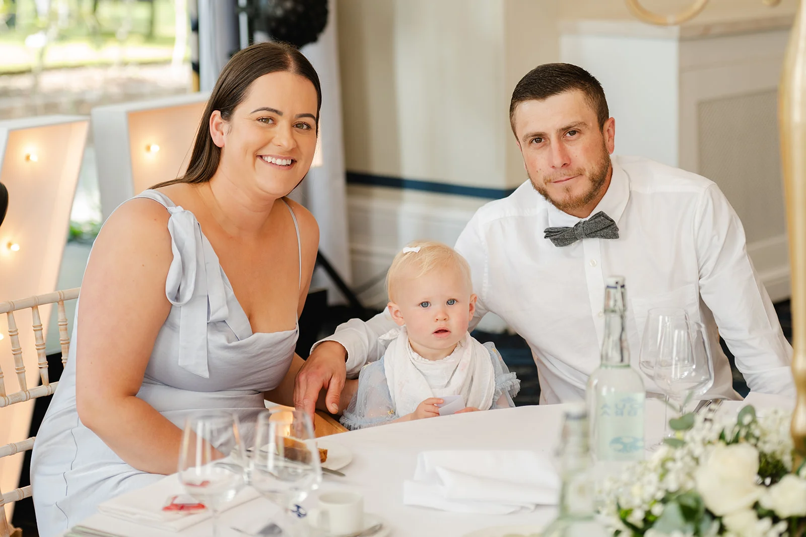 A smiling woman, a groom wearing a white shirt and grey bow tie, and a young baby sit together at a wedding reception table.