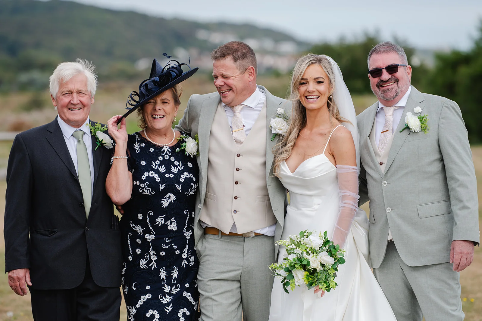 A smiling bride holding a white rose bouquet poses alongside her new husband, parents, and a guest outdoors against a rolling hillside backdrop.