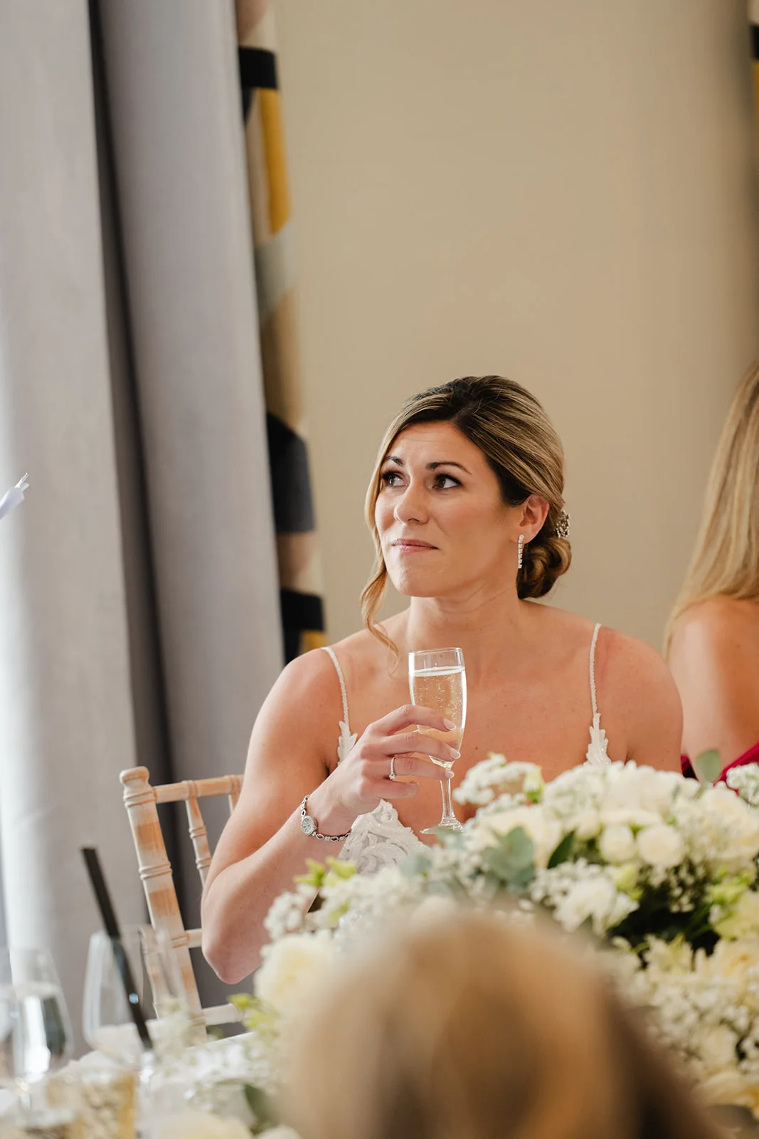 A smiling bride listens to wedding speeches at a reception table while holding a glass of champagne during the wedding breakfast.