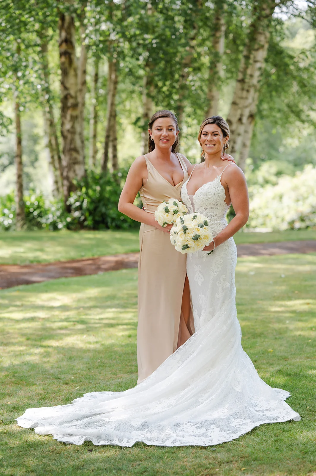 A smiling bride in a lace fit-and-flare wedding dress with a long train stands outdoors in a wooded garden next to her bridesmaid wearing a champagne satin dress, both holding white rose bouquets.