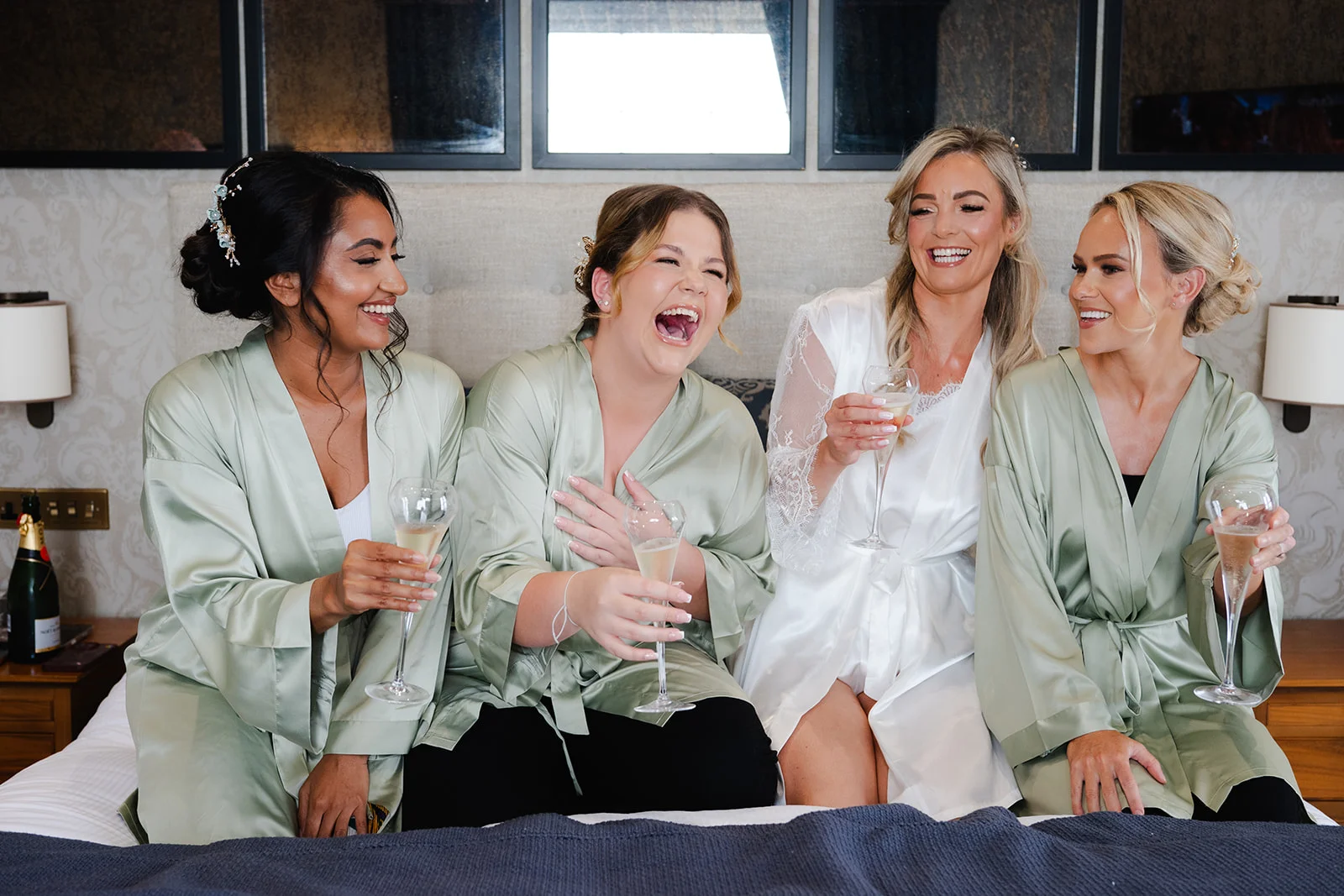 A joyous bride in a white robe and her bridesmaids in matching sage green satin robes sit on a bed while laughing and holding champagne flutes during morning preparations.