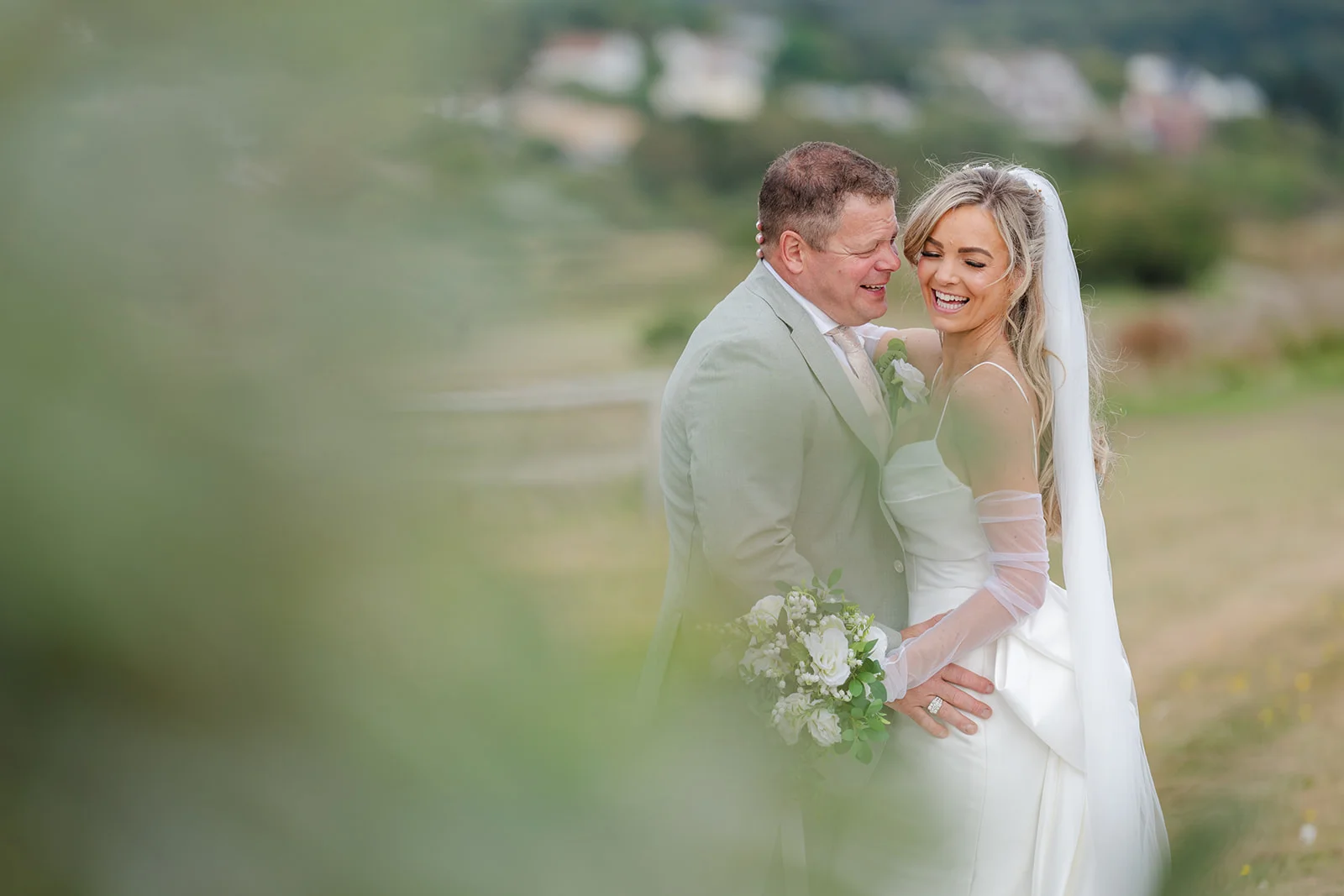 A happy bride and groom laugh together in an outdoor setting framed by soft green foliage, with the bride holding a white bouquet and wearing a sleek satin gown and sheer gloves.