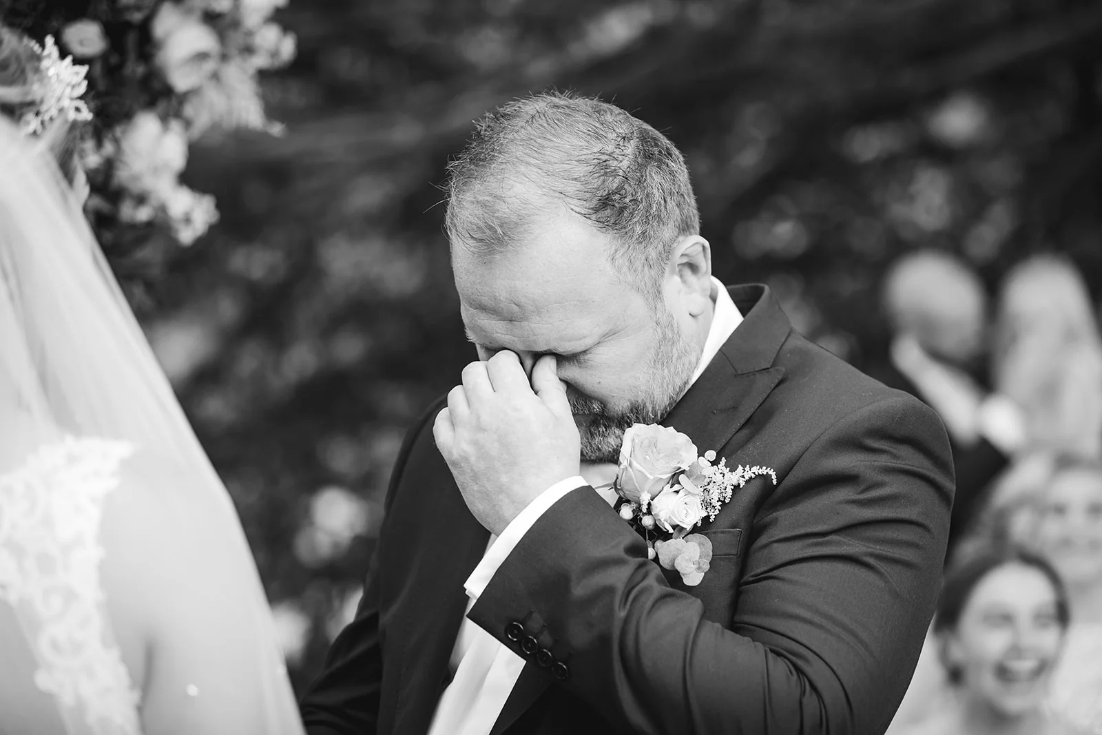 An emotional groom wipes away tears of joy during the wedding ceremony while wearing a dark suit with a floral buttonhole.