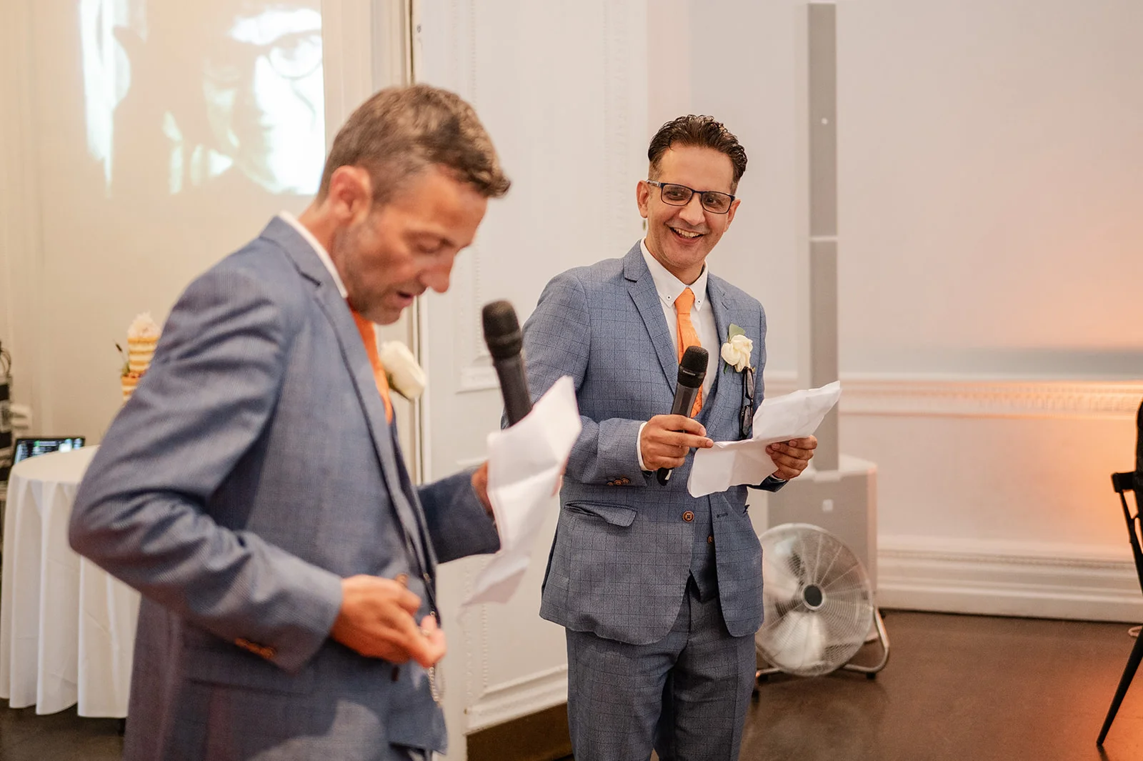 Two smiling grooms in matching blue checked suits deliver wedding speeches holding microphones and notes during the evening reception.