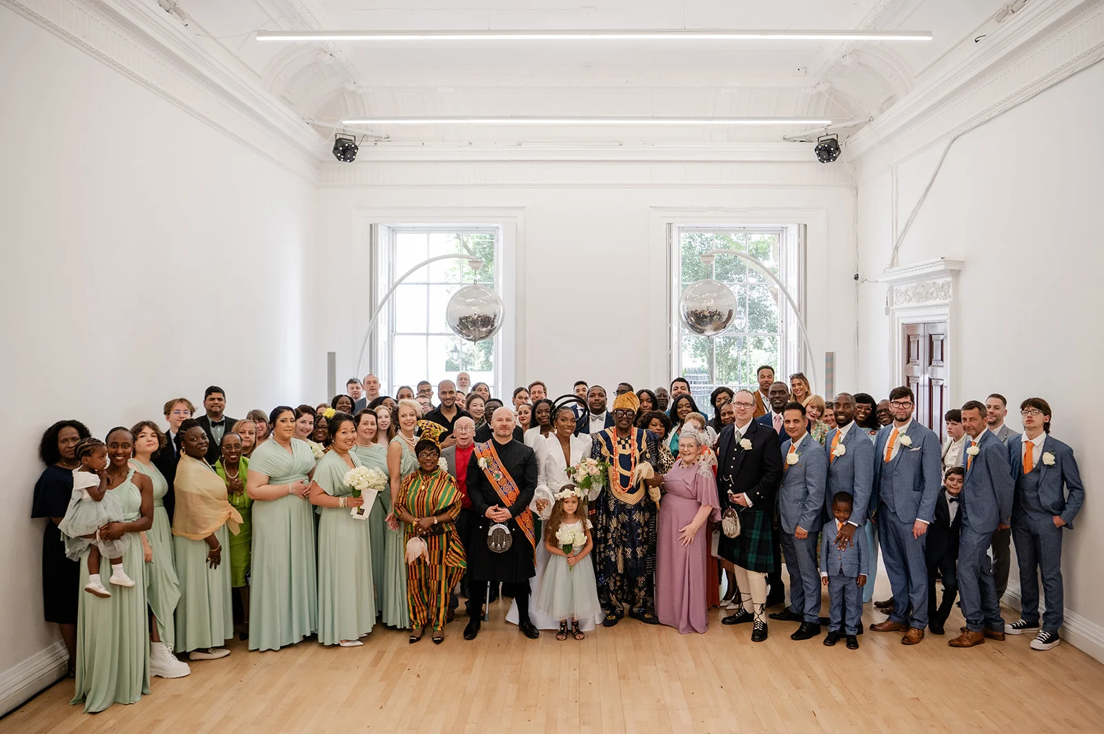 A large wedding group poses for a formal portrait inside a bright, high-ceilinged room with light wooden floors, showcasing diverse guests in coordinated green gowns, blue suits, and traditional cultural attire.