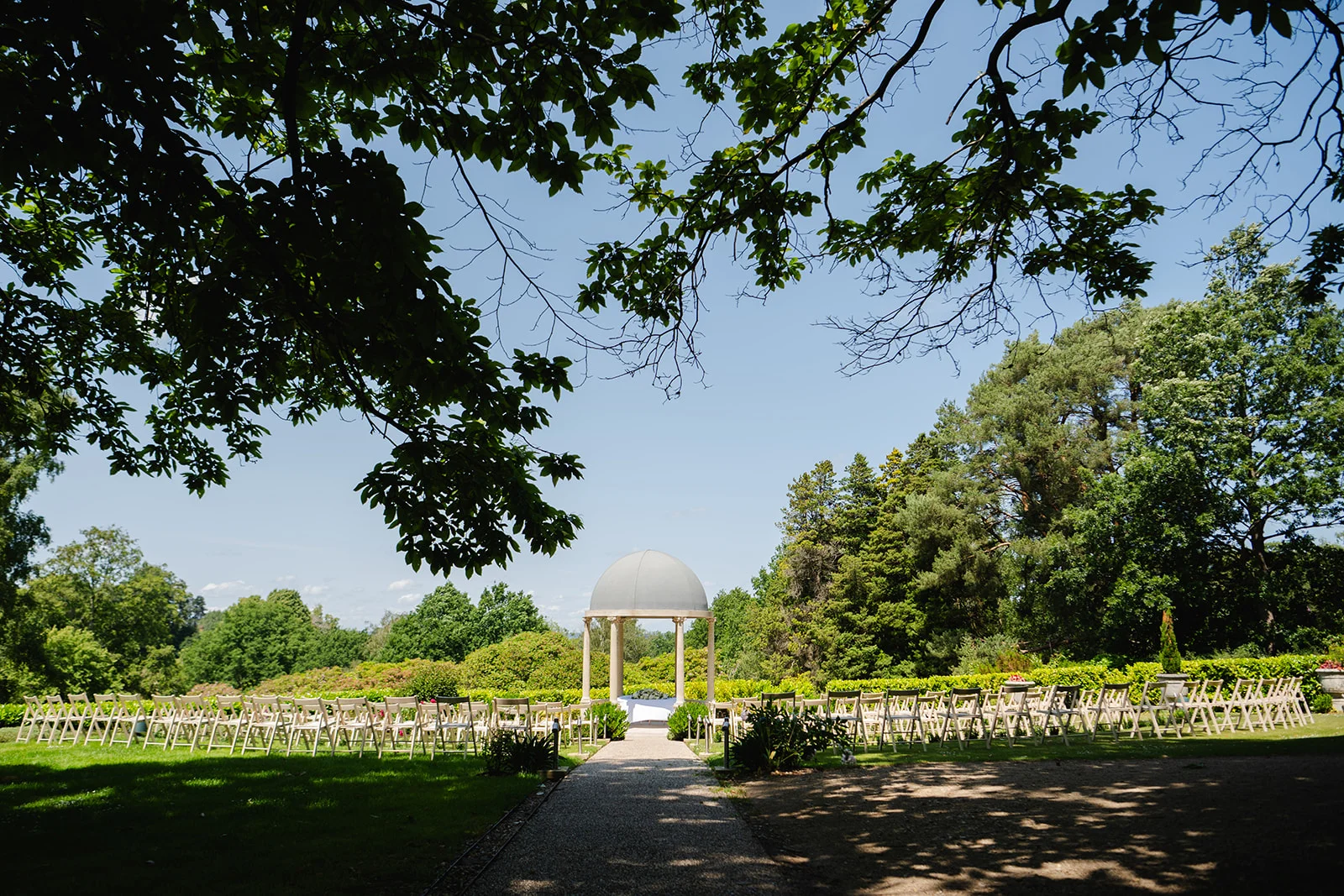 An outdoor wedding ceremony setup framed by overhanging tree branches, featuring a stone gazebo at the end of a gravel aisle lined with white chairs.