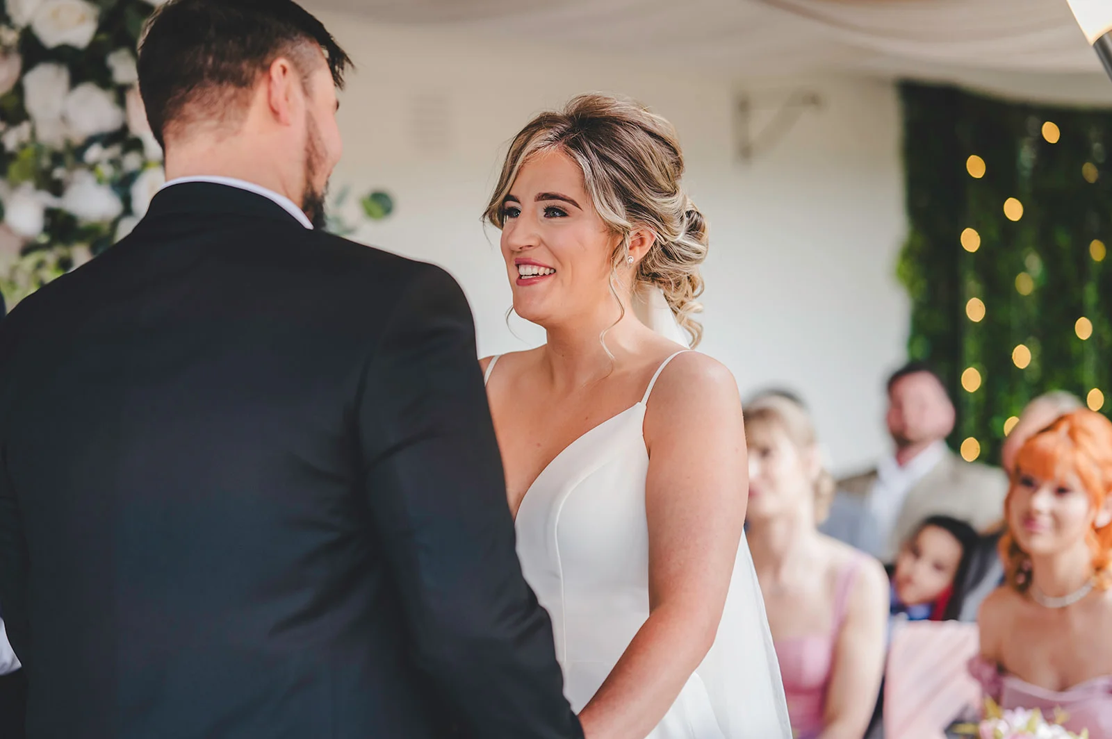 A smiling bride wearing a simple spaghetti strap gown looks lovingly at her groom during the wedding ceremony as guests watch on.